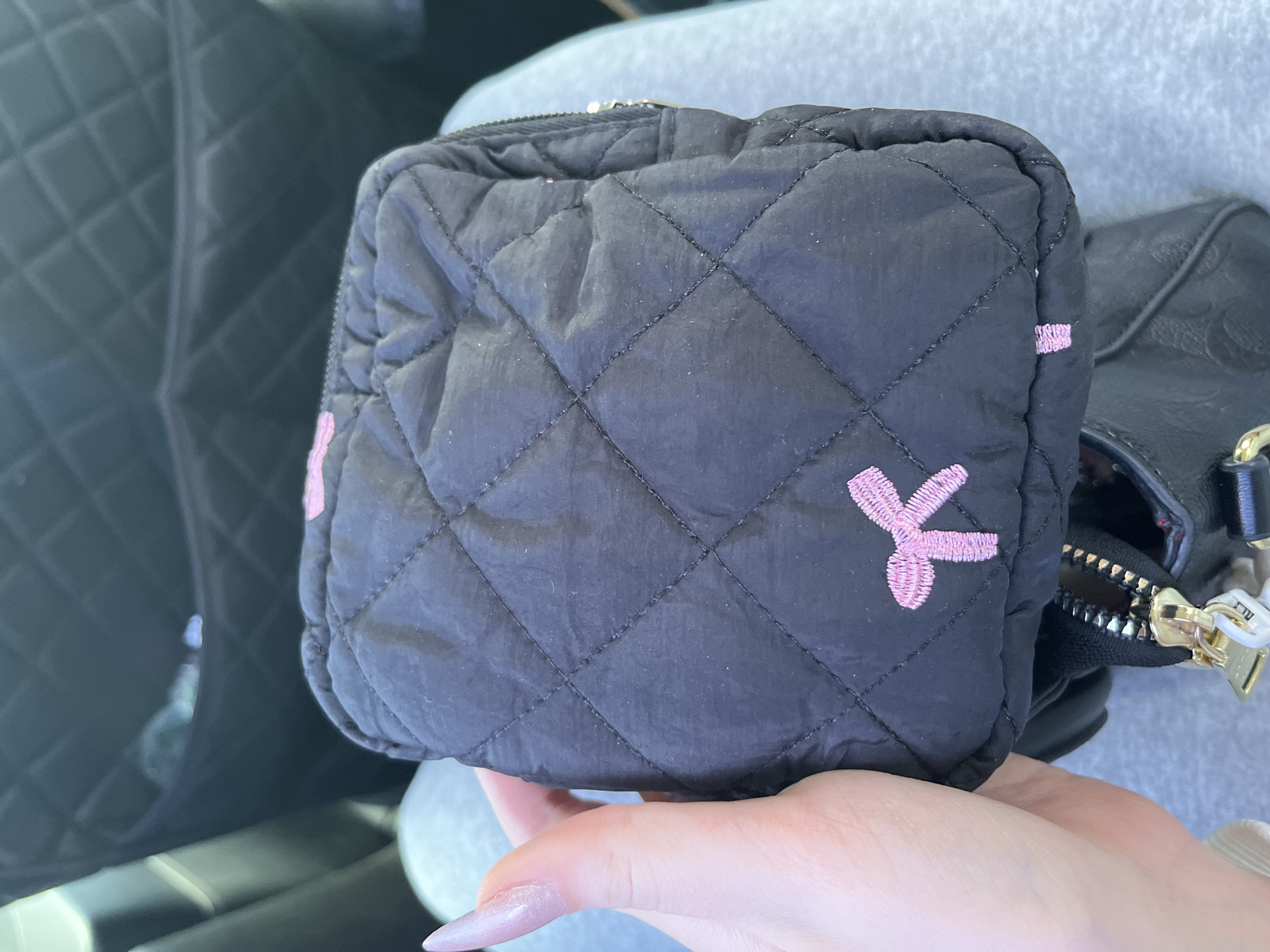 A person's hand with a light pink, long, acrylic fingernail is holding a small, black, quilted makeup bag. The bag has a diamond-pattern quilting with black stitching and pink embroidered details, including a bow on the front and two small rectangular shapes on the bottom. The bag is partially unzipped, revealing a golden zipper pull. The background is out of focus, but appears to be the interior of a car, with quilted black upholstery on the seatback behind the bag, and a person's grey sweatpants visible below. The scene is indoors, and there is no discernible location in the city of Подлесное, Russia.