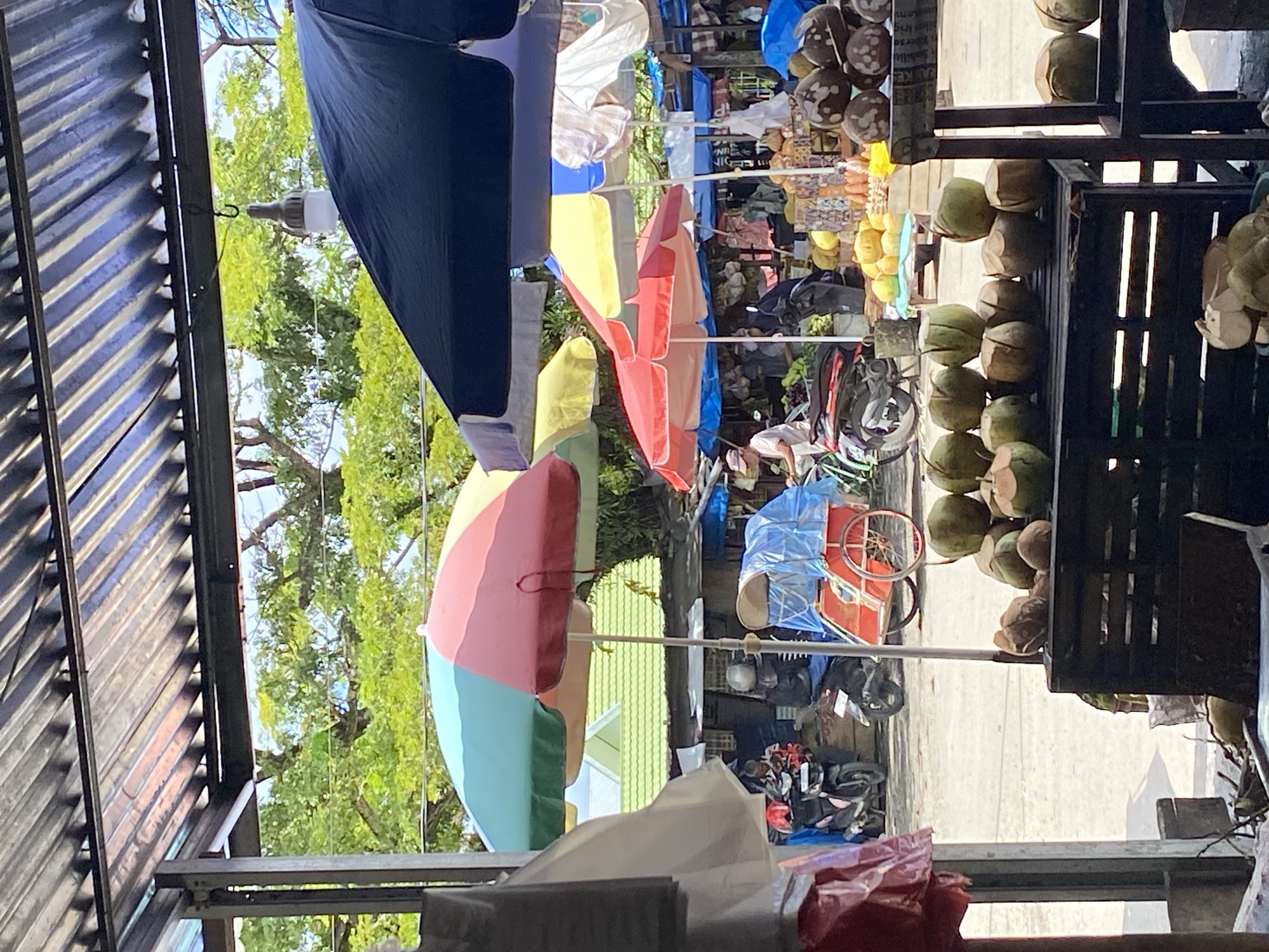 This image depicts an outdoor market scene in Ta', Indonesia. The foreground features a display of coconuts on wooden crates. In the middle ground, various market stalls are visible under colorful umbrellas, with people browsing and interacting. Several motorcycles are parked in the background, suggesting this is a common mode of transportation in the area. The overall environment appears bustling and vibrant, indicative of a local marketplace.