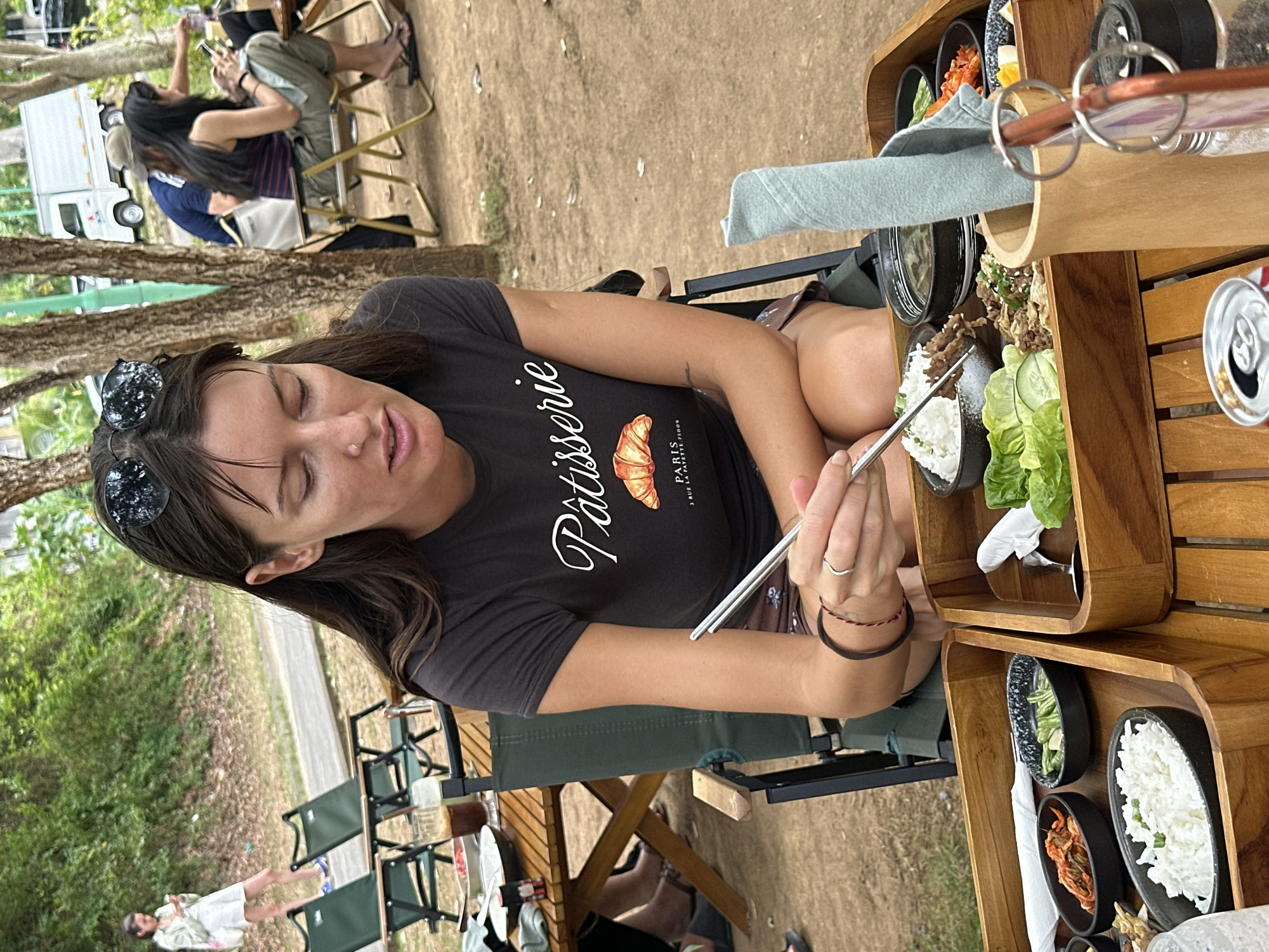 In Kuta Selatan, Indonesia, a woman sits outdoors at a wooden table, holding chopsticks and picking up food. She wears a dark t-shirt with a graphic print, shorts, and sunglasses perched on her head. In front of her, a wooden tray holds several bowls of food, including rice, kimchi, meat, and salad. Another wooden tray is visible to the left. In the background, trees and other people are present, with chairs and a white truck visible. The ground is covered in dirt and grass.