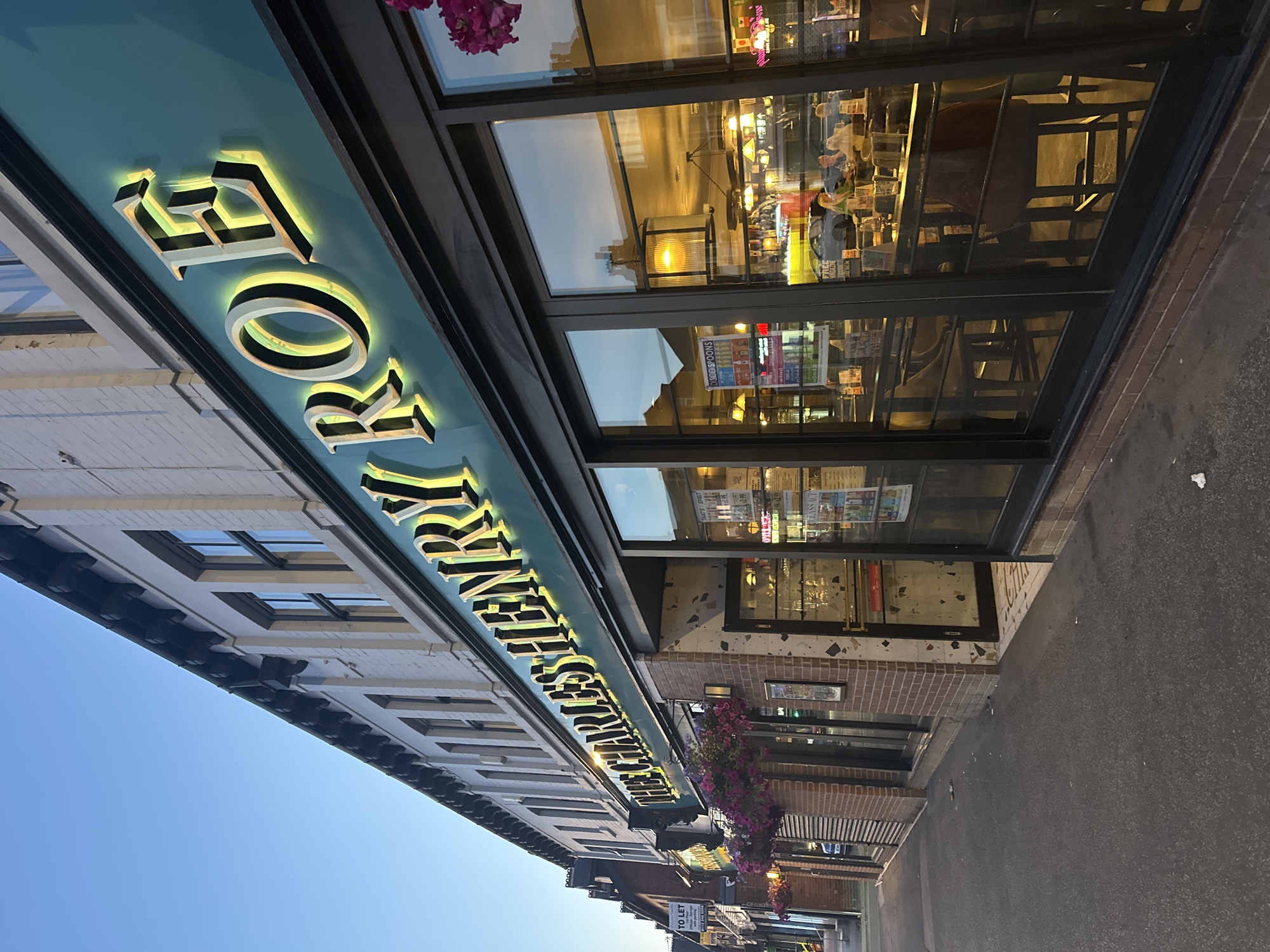 The image shows the exterior of a pub named "THE CHARLES HENRY ROE" located on a street in Leeds, United Kingdom. The pub has a teal-colored facade with illuminated, gold-colored lettering. The building features large glass windows that offer a view into the interior, where tables, chairs, and patrons are visible. Hanging baskets filled with pink flowers are mounted above the entrance. The street in front of the pub is paved, and there are some scattered pieces of litter. The sky above is clear and blue, suggesting it is either early morning or late evening. The building itself appears to be part of a row of similar establishments, with a clear blue sky above. The overall scene conveys a sense of a commercial street in a British city.