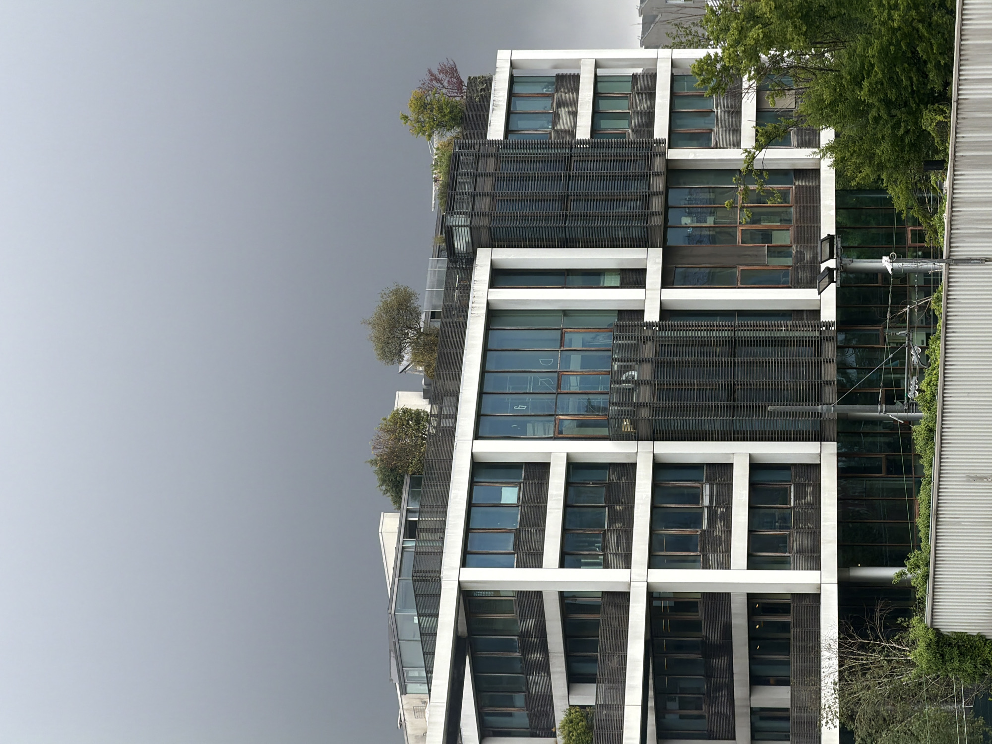 The image captures a portion of a modern architectural building in Milan, Italy, characterized by its geometric facade of white beams and large windows, with some sections covered by wooden slats, suggesting a possible sun-shading system. Trees and other vegetation are visible on some of the building's levels, indicating the presence of planted terraces or rooftop gardens. The sky is overcast, which creates a muted light. The view is a low-angle shot.