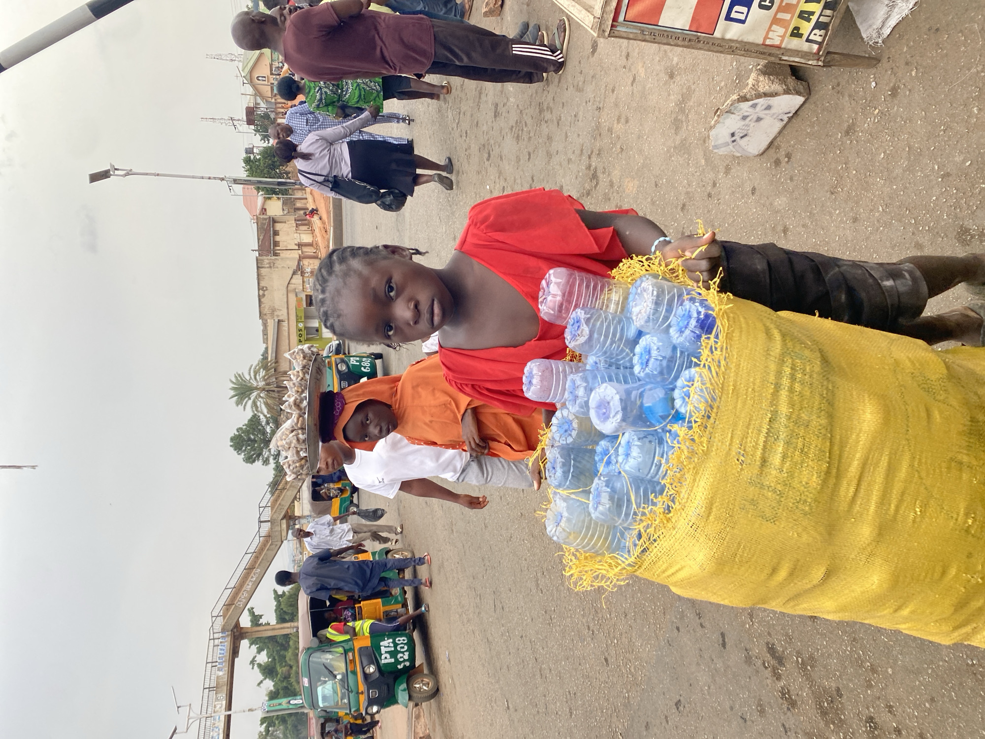 A young girl in a red dress carries a large yellow sack filled with plastic water bottles. She is standing in what appears to be a busy street in Gura Top, Nigeria. In the background, there are other people walking and a three-wheeled vehicle marked "PTA S208". A pedestrian bridge is visible in the distance. The weather appears overcast.