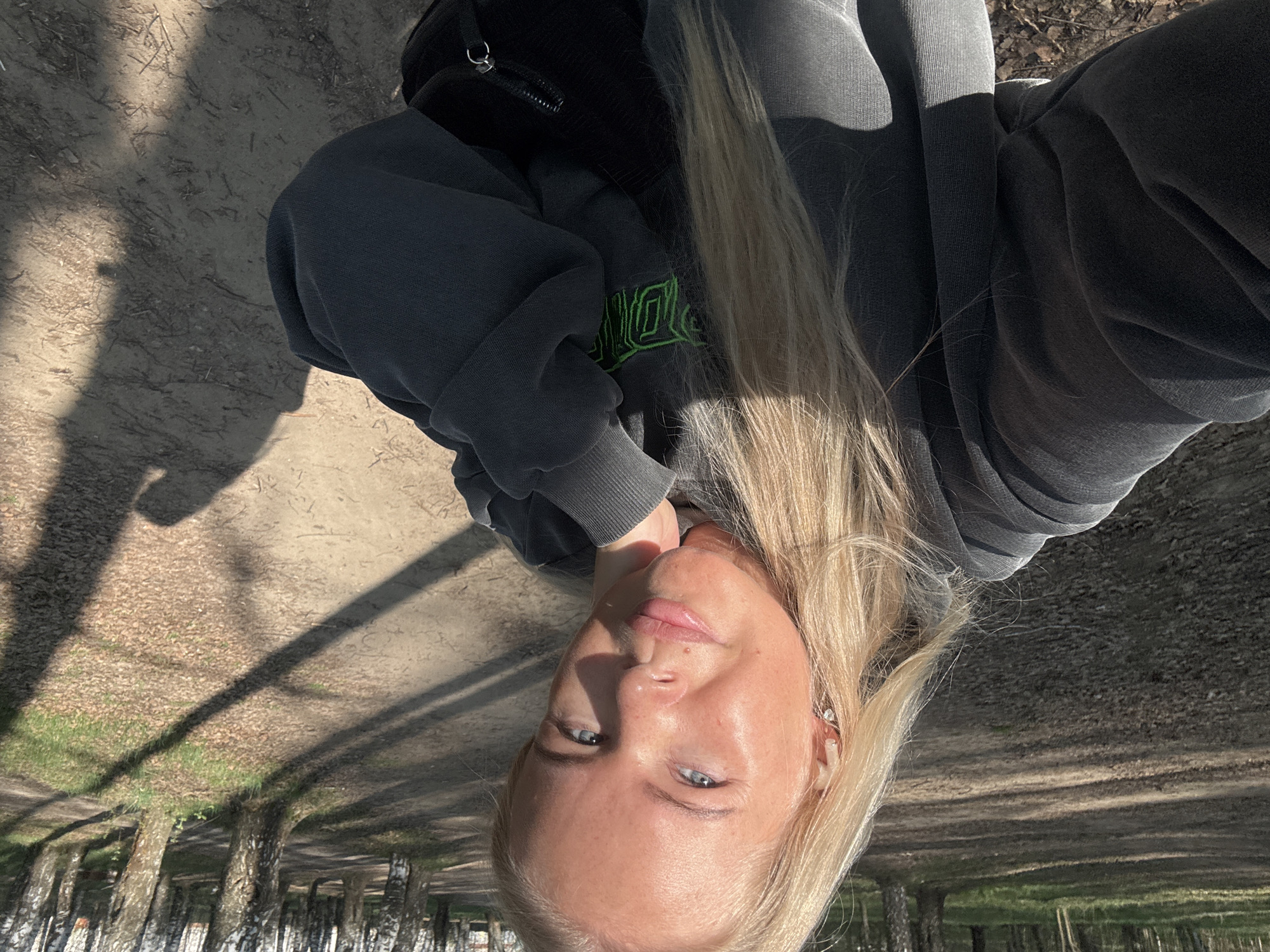 A fair-skinned woman with blonde hair and blue eyes is taking a selfie outdoors. She is wearing a gray hoodie with green writing on the front and a black bag slung over her shoulder. The background shows a path lined with trees casting long shadows. The location is Chaikovsky, Russia, suggested by the birch tree species on the side of the road.