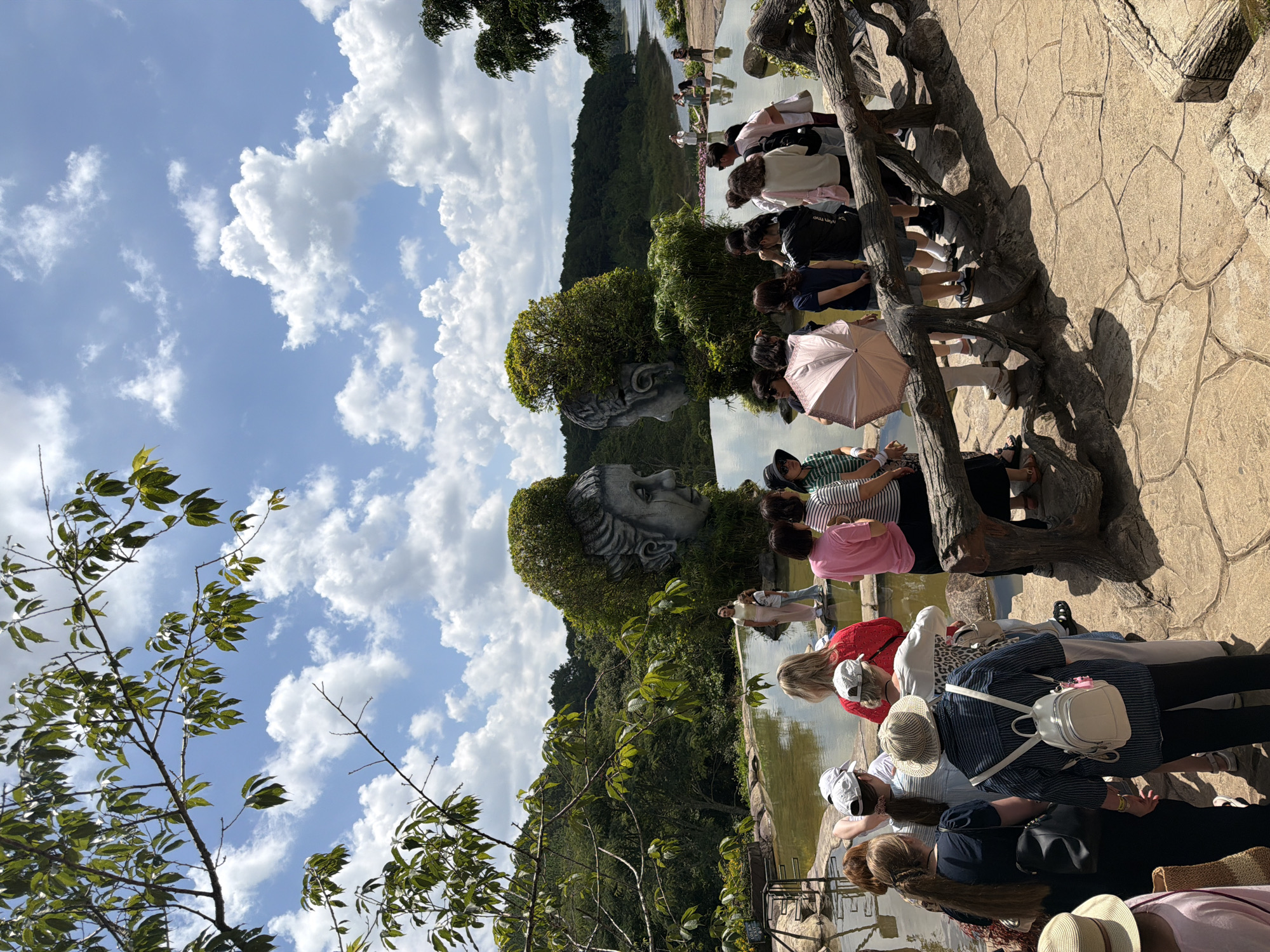 In Da Lat, Vietnam, a group of tourists gathers around a water feature adorned with two large, moss-covered sculptures of human heads facing each other. The sculptures, reminiscent of ancient busts, are integrated into the lush greenery surrounding a calm body of water. Several individuals are captured mid-action, some posing for photos, others observing the unique art installation, and a few conversing amongst themselves. The foreground features a textured, stone pathway and a rustic wooden railing, suggesting a well-maintained tourist attraction. The background reveals a dense forest and a tranquil body of water stretching towards the horizon under a sky dotted with fluffy white clouds. The overall scene depicts a serene and picturesque landscape, characteristic of the natural beauty and artistic flair found in Da Lat.