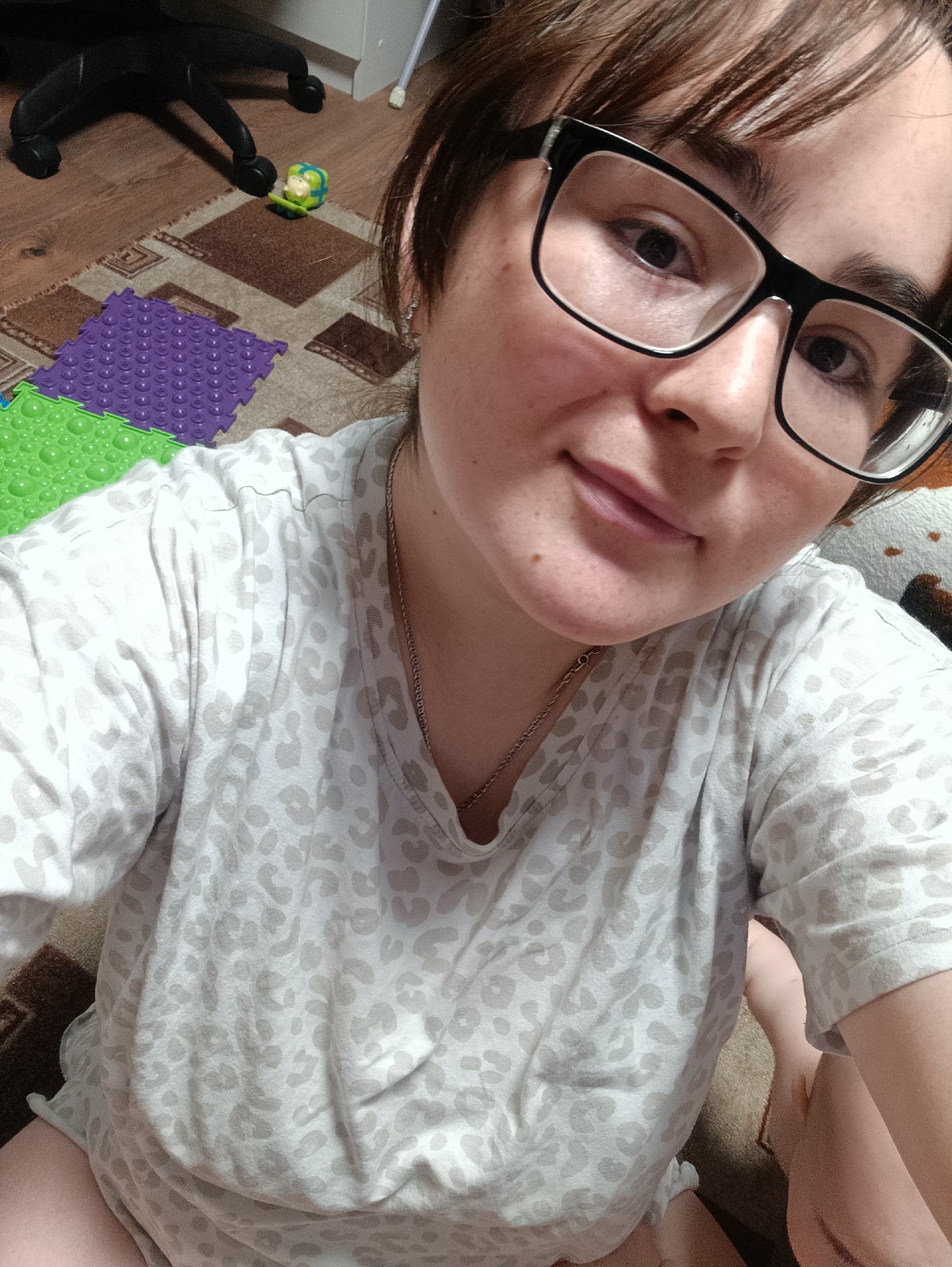 An indoor scene depicts a female subject in a close-up, selfie-style photograph. The subject has dark hair, dark-framed eyeglasses, and is looking directly at the camera with her lips slightly upturned. She is wearing a light-colored t-shirt with a grey leopard print pattern and a thin chain necklace.
The background shows a wooden plank floor partially covered by a brown patterned rug. On the rug, two interlocking textured floor mats, one green and one purple, are visible. A small green and blue toy figurine is positioned on the rug near a partially visible black office chair base. A white baseboard or wall section is present in the upper right background. The exact location within the city of Noviy, Russia, cannot be determined from the provided image data; however, the setting is consistent with a residential interior.
