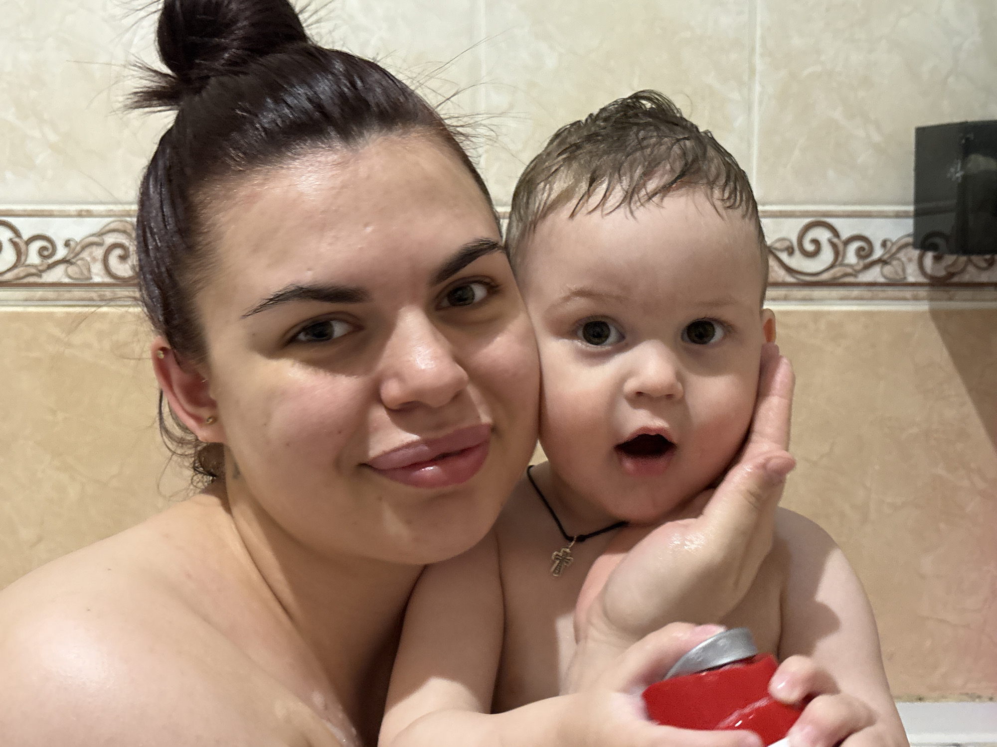 A woman and a baby are in a bathtub. The woman has dark hair pulled into a bun and is smiling at the camera. The baby has wet hair, a silver cross necklace, and is holding a red object. They are in a bathroom with beige and brown patterned tiles. The location is Unknown, Unknown.