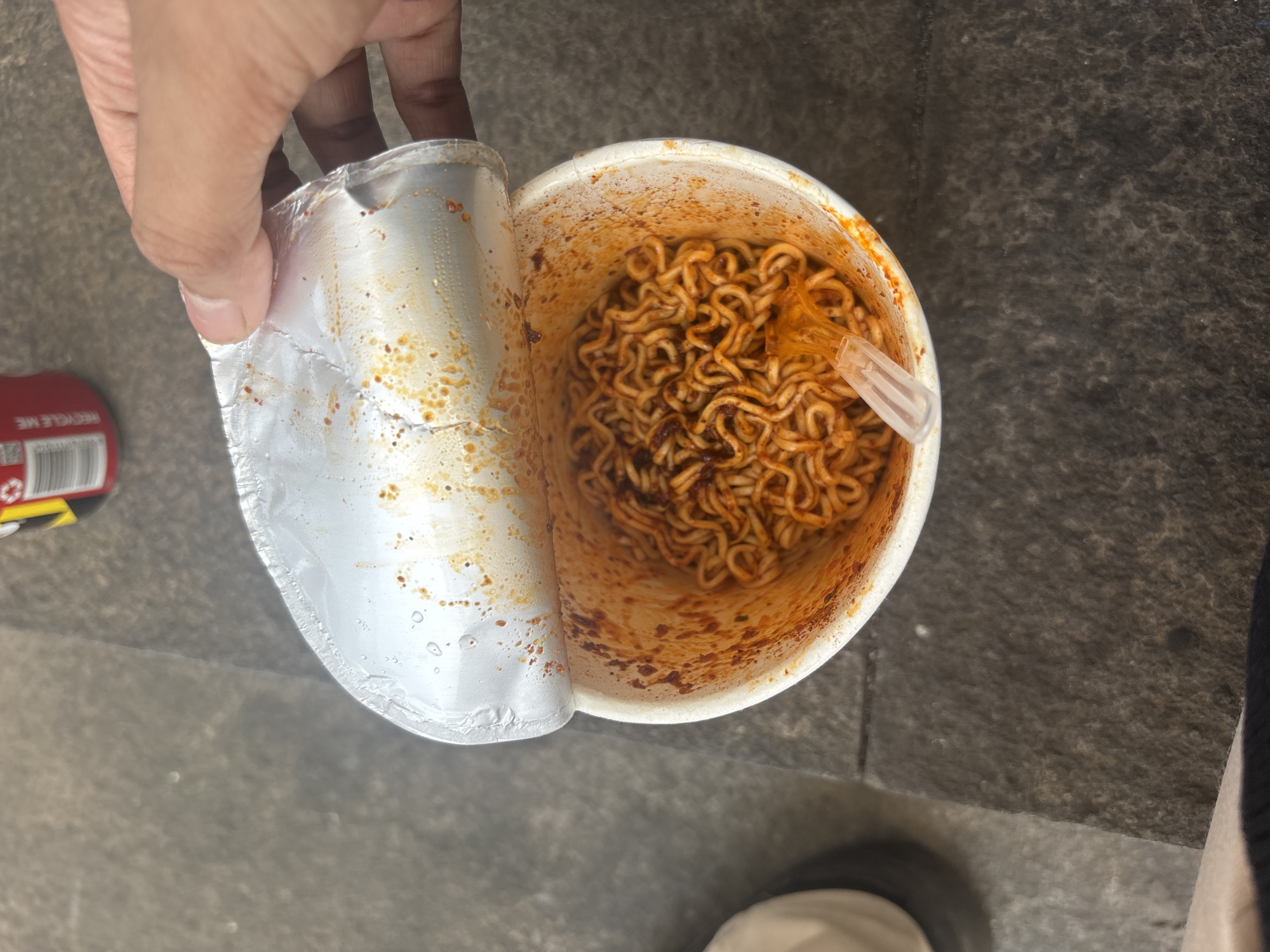An overhead shot shows a person's hand peeling open a foil lid from a cup of instant noodles. The noodles are coiled tightly within the white cup, coated in a red, spicy sauce. A clear plastic fork rests in the noodles. In the background, a portion of a red and yellow can is visible. The noodles and cup are placed on a textured grey surface, possibly pavement or a stone countertop. The setting is a city street in North Jakarta, Indonesia.