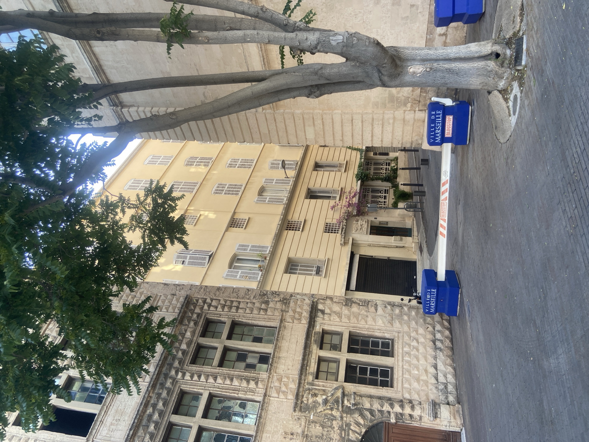 A street in an urban environment is depicted. The street is paved with dark grey cobblestones. On the left side of the image, a stone building with ornate detailing and mullioned windows stands. To the right of this building, a lighter colored, multi-story building with numerous windows and white shutters is visible. A large tree with a textured trunk and several branches extends from the right side of the image, obscuring parts of the buildings. In the foreground, a white barrier with orange and white diagonal stripes is positioned across the street, supported by two blue, rectangular blocks. One of the blue blocks has the text "VILLE DE MARSEILLE" and "BLOCSTOP" printed on it in white lettering. Another blue block is partially visible on the far right side of the image. The scene appears to be a street closure or controlled access point within the city of Marseille, France. The lighting suggests it is daytime, with sunlight illuminating the buildings and casting shadows.