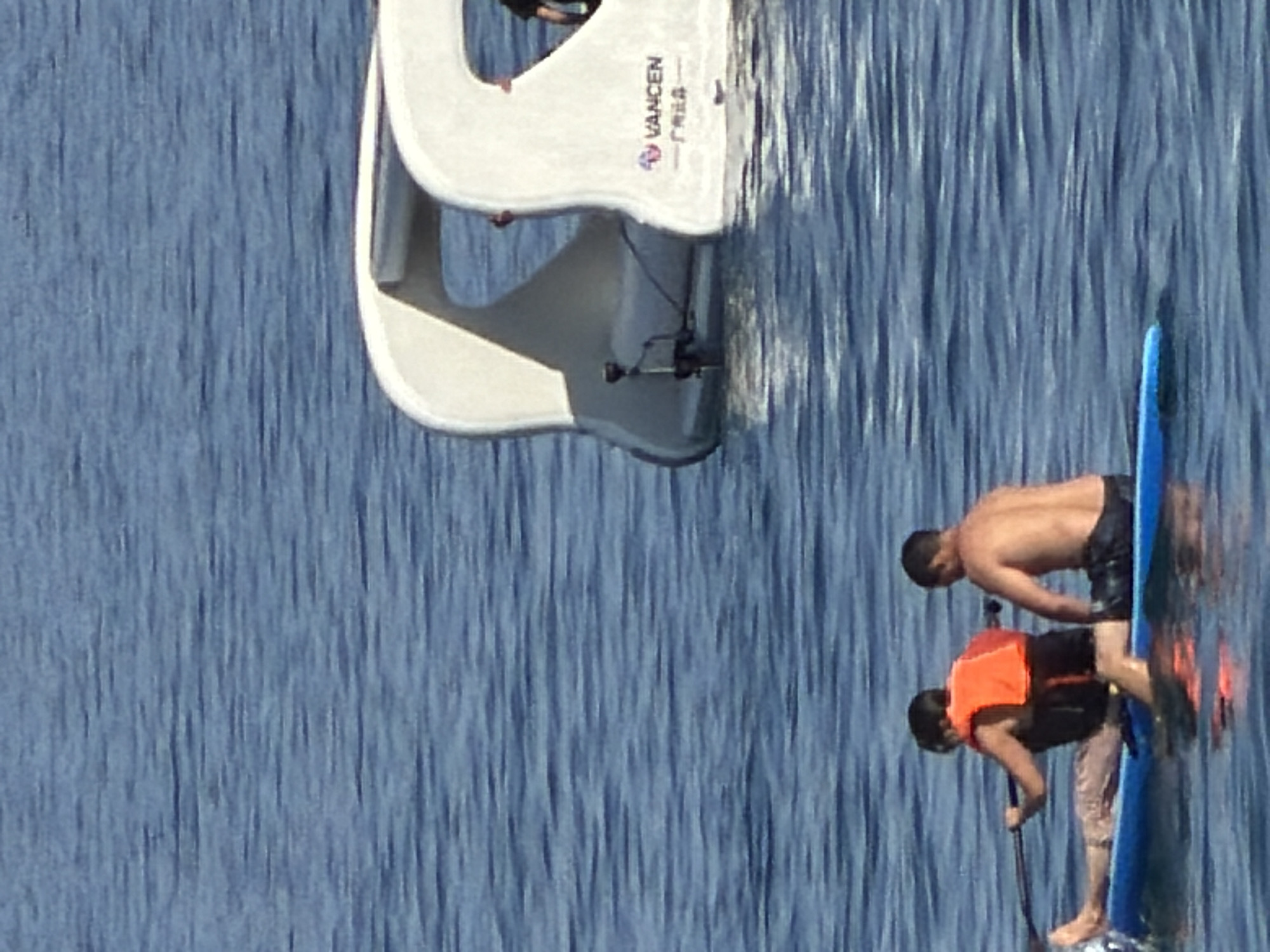 Two individuals are on a paddleboard on the water. The person in the foreground on the left is wearing an orange life vest and is paddling. The person behind them on the right is shirtless and appears to be sitting. In the background, a white, futuristic-looking boat or structure is partially submerged in the water. The water is blue and appears calm with ripples.