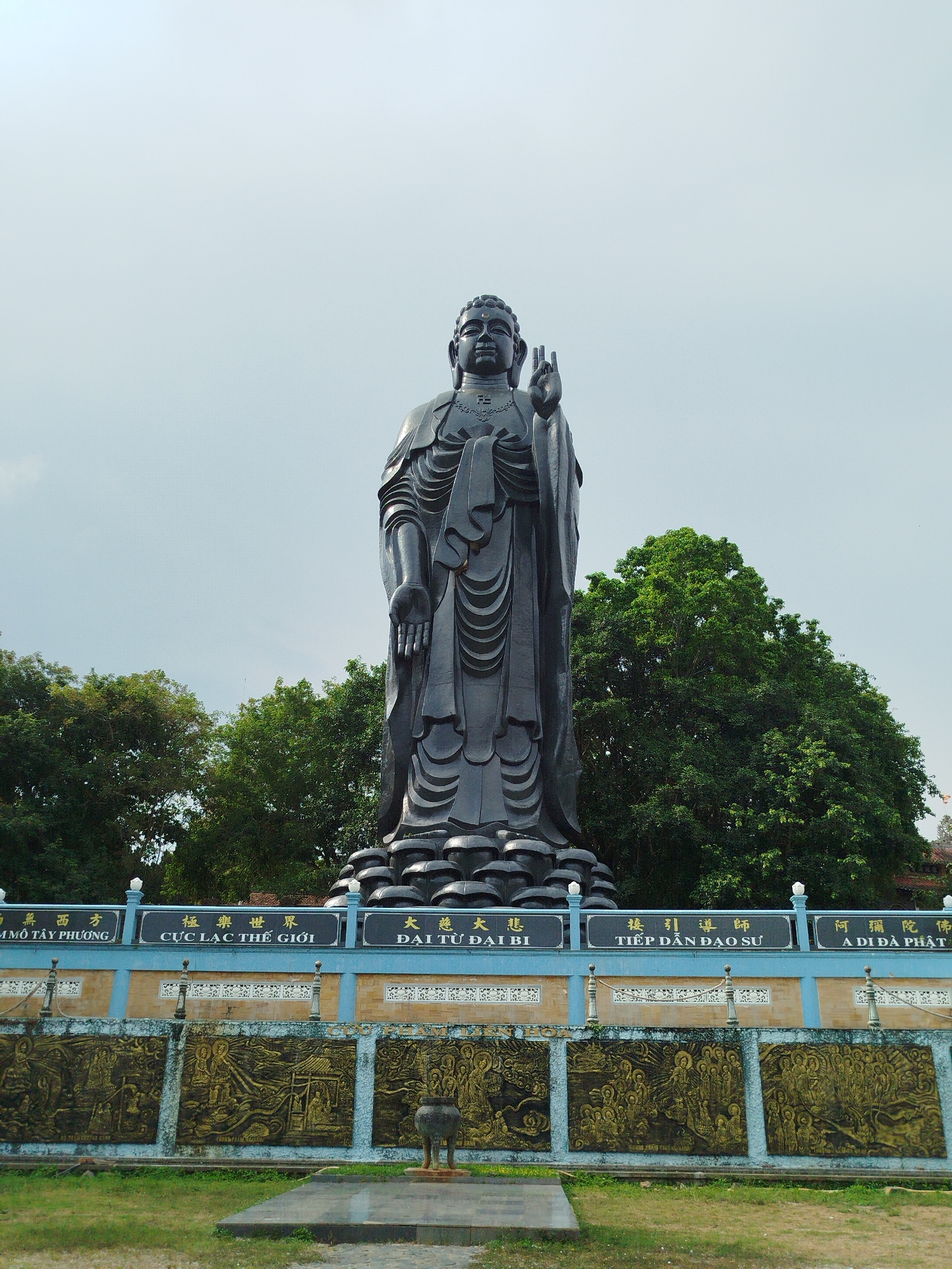 The image shows a large, dark-colored statue of a standing Buddha figure, likely made of stone or metal. The statue is positioned in front of a wall with inscriptions in Asian characters and relief carvings. The inscription board has Chinese and Vietnamese text. Trees are visible in the background. A platform and a small incense burner are located at the base of the statue. The location is Phú An, Vietnam.