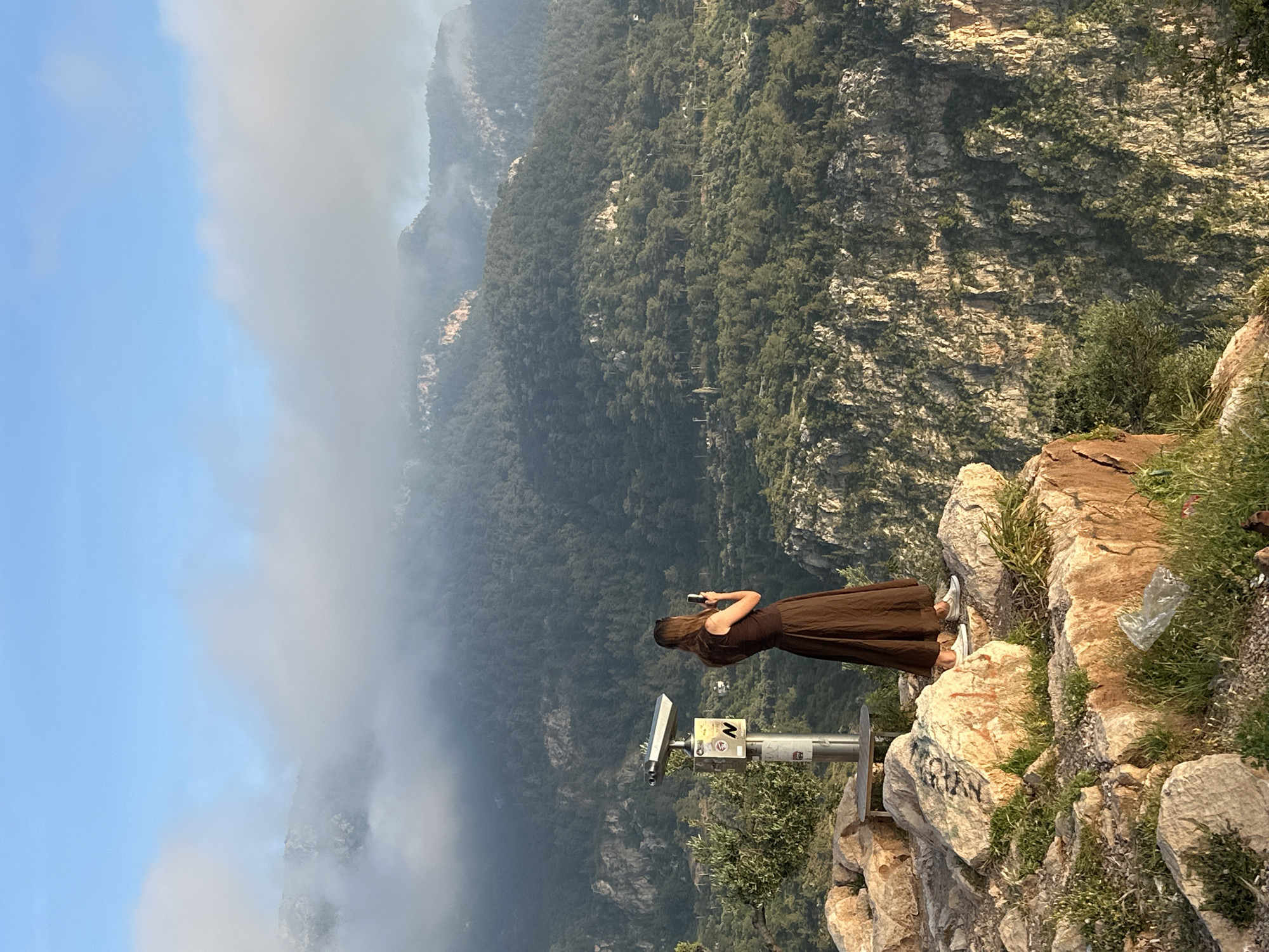 The image captures a scenic mountainous landscape from a high viewpoint, likely a tourist overlook in Fethiye, Turkey, during daytime.
In the foreground, a person is seen from behind, standing on a rocky ledge. They wear a long, dark brown dress and white sneakers, holding a mobile phone up as if taking a photo or video of the expansive view. To their left, a coin-operated viewing telescope with a silver-grey body and stand is positioned, oriented towards the distant mountains. The stand of the telescope has various stickers, including a prominent "N" (likely indicating North) in a white box. The ground immediately around the person and telescope consists of light-colored rocks, sparse green vegetation, and a discarded clear plastic bag on the right. Faint, dark graffiti, possibly "NGEL", is visible on a rock in the lower foreground.
The background reveals a vast panorama of rugged, layered mountains covered in dense green forests. Valleys wind through the terrain, with hints of distant settlements. The far-off landscape is partially obscured by a soft, light haze or clouds that blend with the blue sky above. The atmospheric conditions create a sense of depth and scale, with the misty distance contrasting with the clear foreground. The scene evokes a sense of natural grandeur and contemplation, typical of a scenic mountain vista.