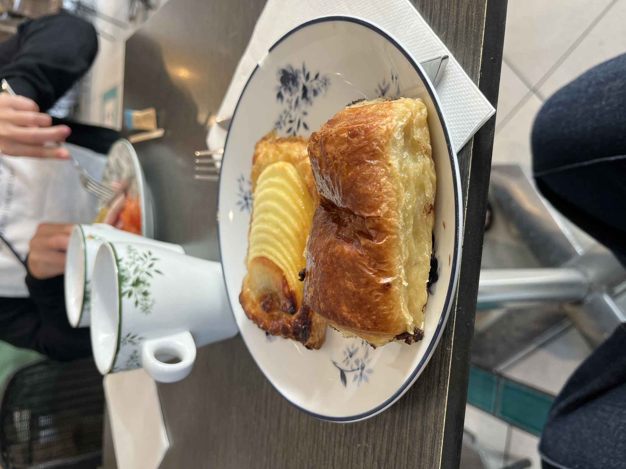 The image captures a close-up view of a plate containing two pastries - one with thinly sliced fruit arranged in a fan pattern, and another appearing as a layered, golden-brown cube. The plate features a white base with a blue floral pattern and a blue rim. A white napkin is positioned near the edge of the plate, and silverware is visible alongside it. Two white cups with green floral decorations are placed near the plate. A person in a dark-colored jacket is seen using silverware on a plate located in the background. Part of another person is visible on the side of the frame. The scene is set on a dark table, with tile flooring observable in the background. No other identifying details are present to confirm the location in Grimaud, France.