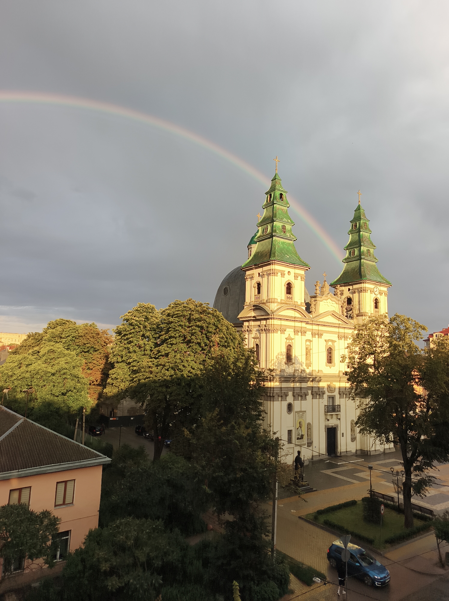 The image depicts a wide shot of Ternopil, Ukraine, showcasing the Church of the Virgin Mary of Perpetual Help. A rainbow arches across the overcast sky, adding a vibrant splash of color to the scene. The church, a grand architectural structure, features two prominent green-topped spires, ornate white facades, and a large dome. In the foreground, lush green trees frame the church, with a few buildings and a street visible in the lower portion of the frame. A blue car is parked on the street, with a person standing nearby. The overall atmosphere suggests a post-rain setting, with the sky still holding onto some clouds.