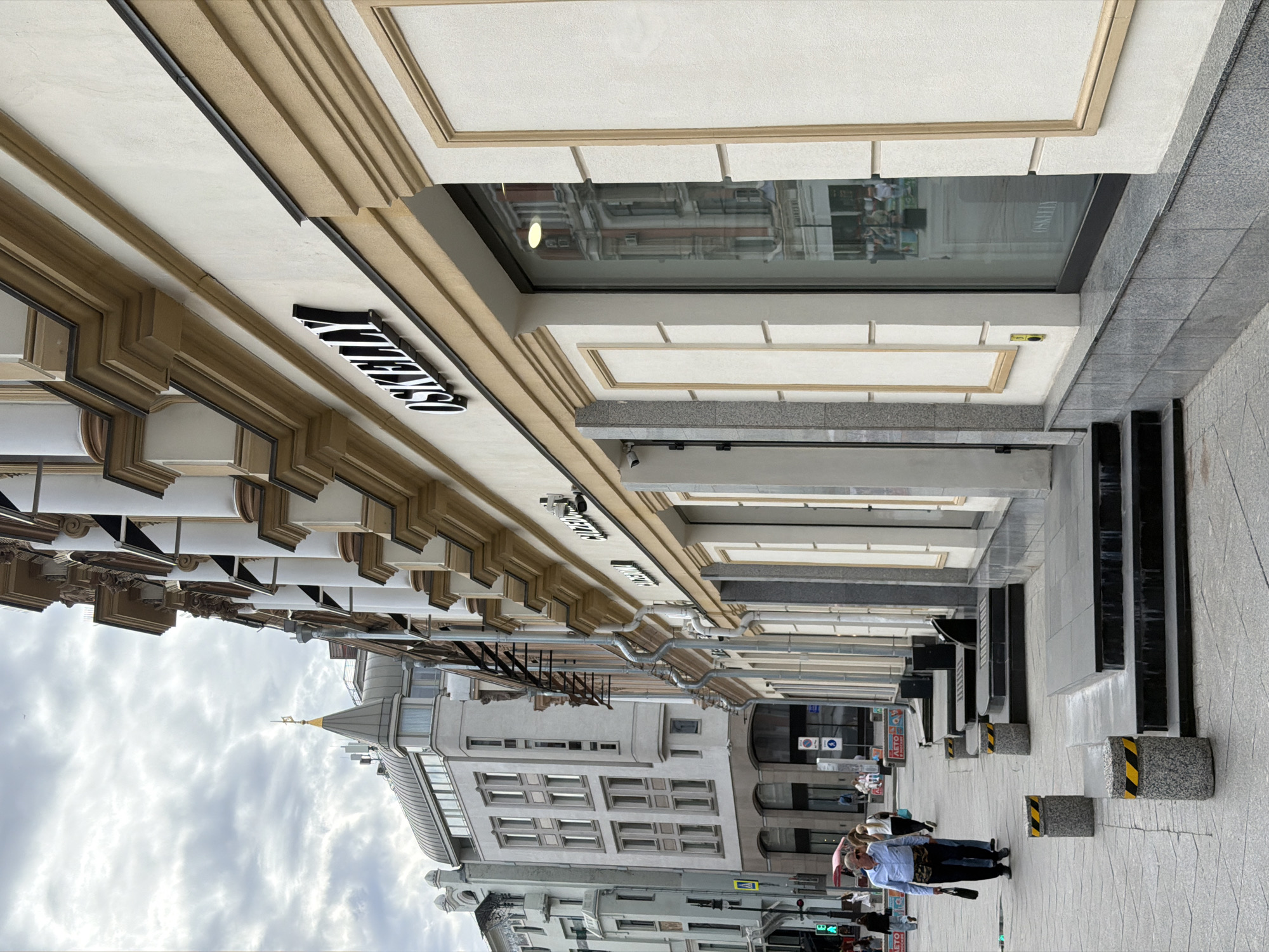 The image depicts a street in Moscow, Russia. The perspective is from a low angle, looking upwards at the facades of buildings lining the street. The architecture is ornate with detailed trim and decorative elements. At least one building has a sign reading "Kuznetsky". The sky is overcast. On the street level, there are pedestrians. The sidewalk is gray and has concrete barriers marked with black and yellow stripes.