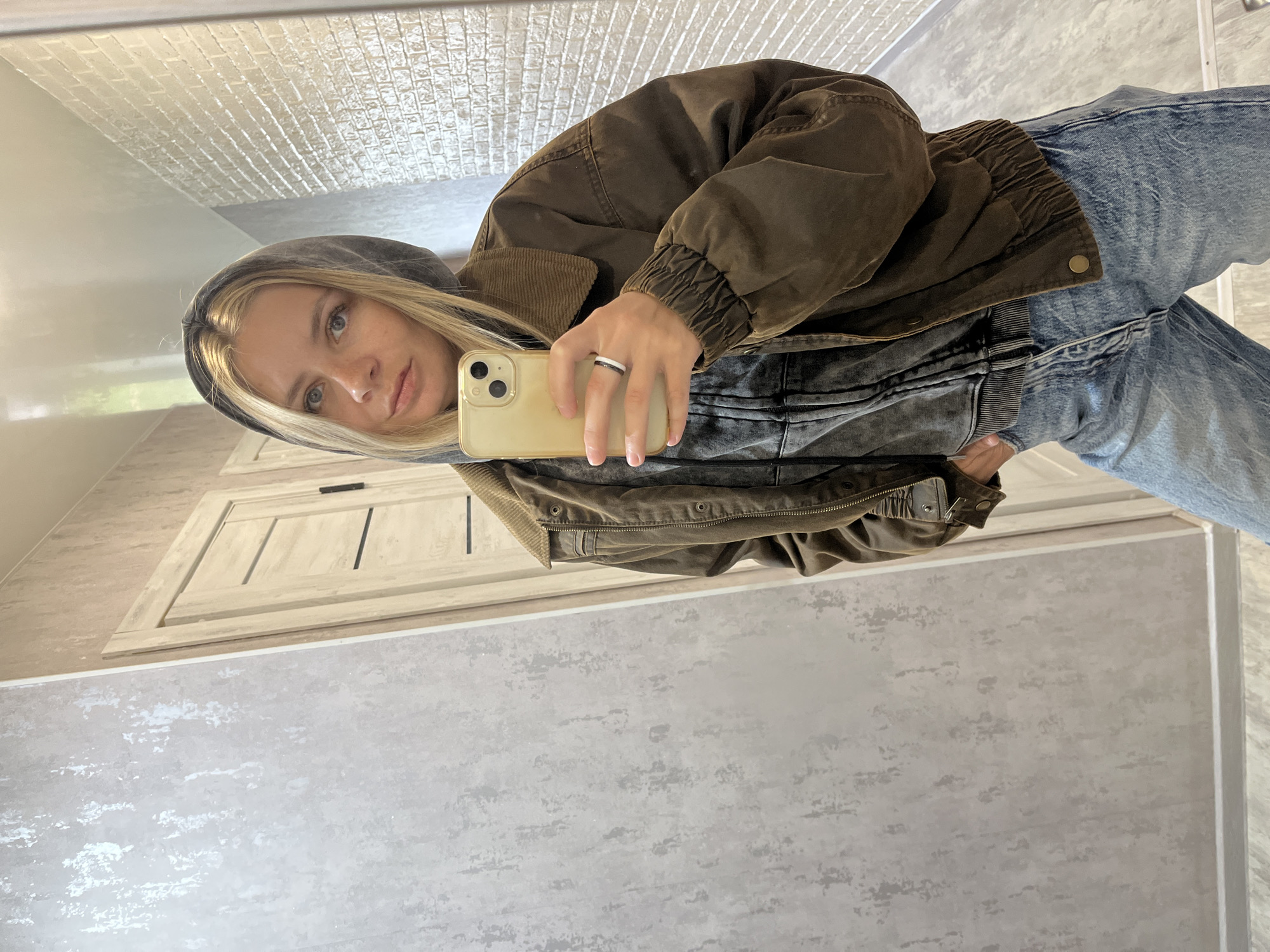 A blond woman in a brown hooded jacket and blue jeans is taking a mirror selfie with a gold iPhone. She is standing indoors with a light-colored door behind her. The location is likely in Chaikovsky, Russia. The floor and wall are neutral-colored. The woman is holding the phone in her left hand, which has a silver ring with a black stripe.