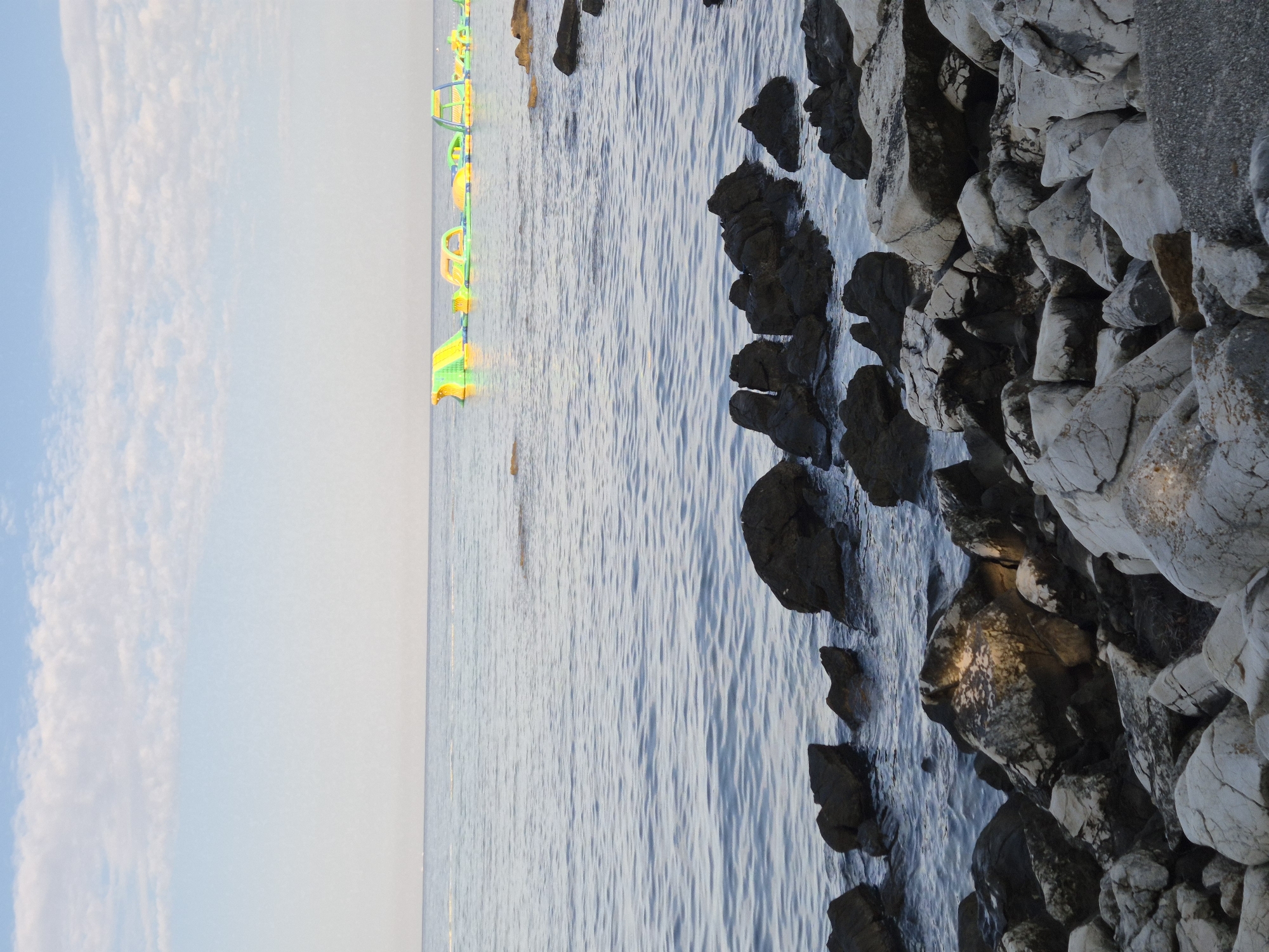 This is a photograph of the Adriatic Sea at Monterol, Croatia. In the foreground, a rocky shoreline with various sizes of grey and white stones is visible. The sea is calm with gentle ripples and a few scattered rocks near the shore. In the distance, a series of inflatable water toys, primarily green and yellow, are floating on the water. The sky is a pale blue with scattered white clouds. The scene appears to be set during daylight, likely late afternoon or early morning due to the soft light. There are no people visible in the image.