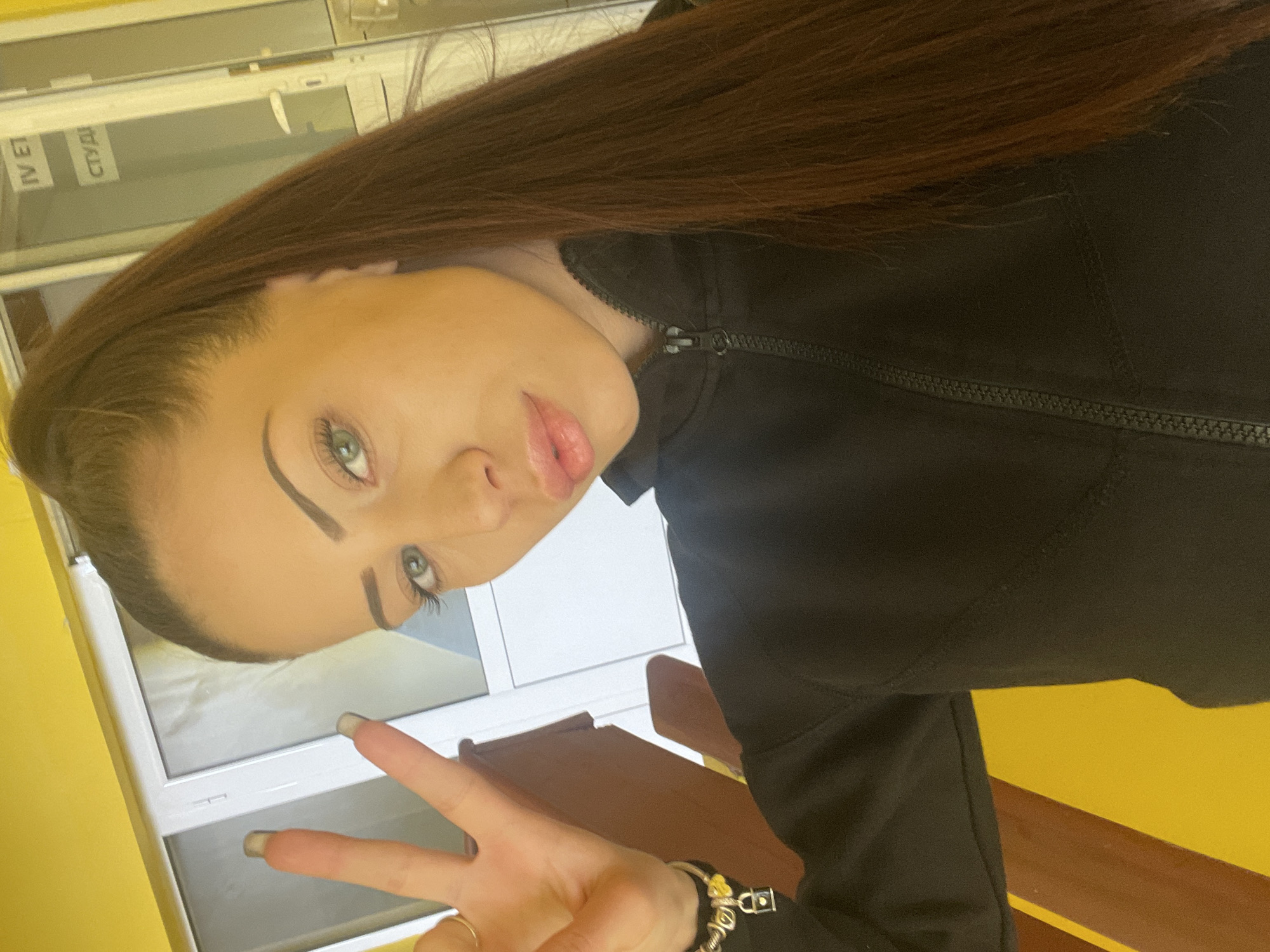 A woman with long, dark brown hair, light skin, and blue eyes is positioned in front of a yellow wall. She is wearing a black zip-up jacket. She is making a peace sign with her right hand. A bracelet is visible on her wrist. Behind her, there is a white-framed structure with what appears to be a window or a display with text in Cyrillic, "IV ET СТУД". The location is suggested to be in Veliko Tarnovo, Bulgaria, although specific details to confirm this are not visually present in the image.
