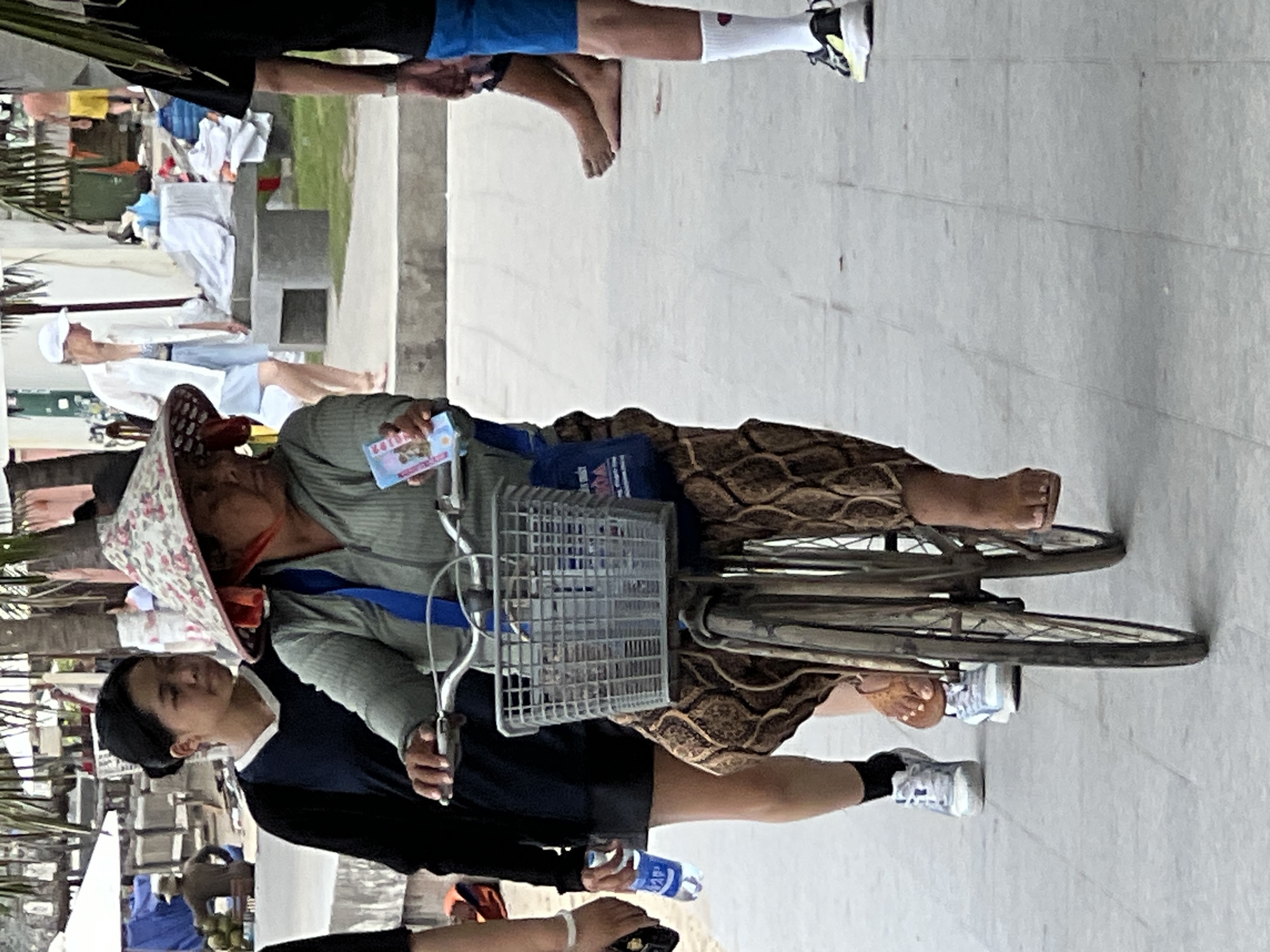 The image depicts a street scene in Nha Trang, Vietnam, likely near a beach or promenade. In the foreground, a woman with a wide-brimmed hat is riding a bicycle. She is wearing a light green long-sleeved shirt and a patterned skirt. Her hands are on the handlebars, and a wire basket is attached to the front of the bicycle, containing items that are not clearly identifiable. The woman is holding a small, colorful card in her right hand. Behind her, to the left, a young woman wearing a dark long-sleeved shirt and white sneakers stands holding a plastic bottle of water. Further in the background, to the right of the cyclist, a person in black shorts and white socks with athletic shoes is visible. To the left of the cyclist, partially obscured, is another individual wearing dark clothing. The environment appears to be an outdoor walkway or street, with palm trees and some structures in the background, suggesting a tropical urban setting. There are also piles of items and what appear to be market stalls in the distance. The overall impression is one of a casual, everyday scene in a Vietnamese coastal city.