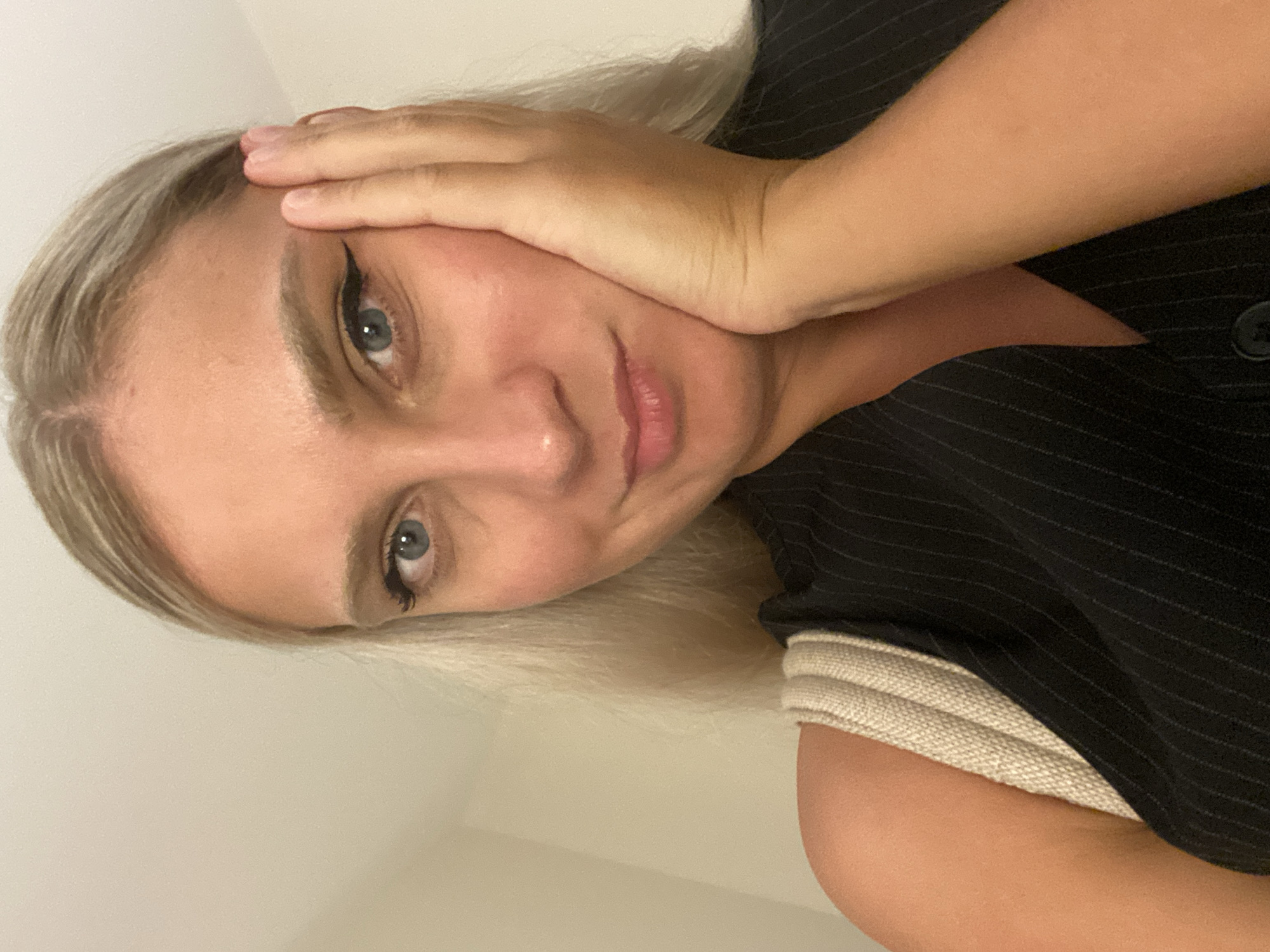 A young Caucasian woman with blonde hair and blue eyes is captured in a close-up selfie. She has on a black pinstripe vest and a light-colored strap is visible over her shoulder. Her right hand is resting on her cheek, and she is looking towards the camera with a slight smile. The background is a plain, light-colored wall, suggesting an indoor setting. There is no indication of the city of Aksu, Turkey within the image.