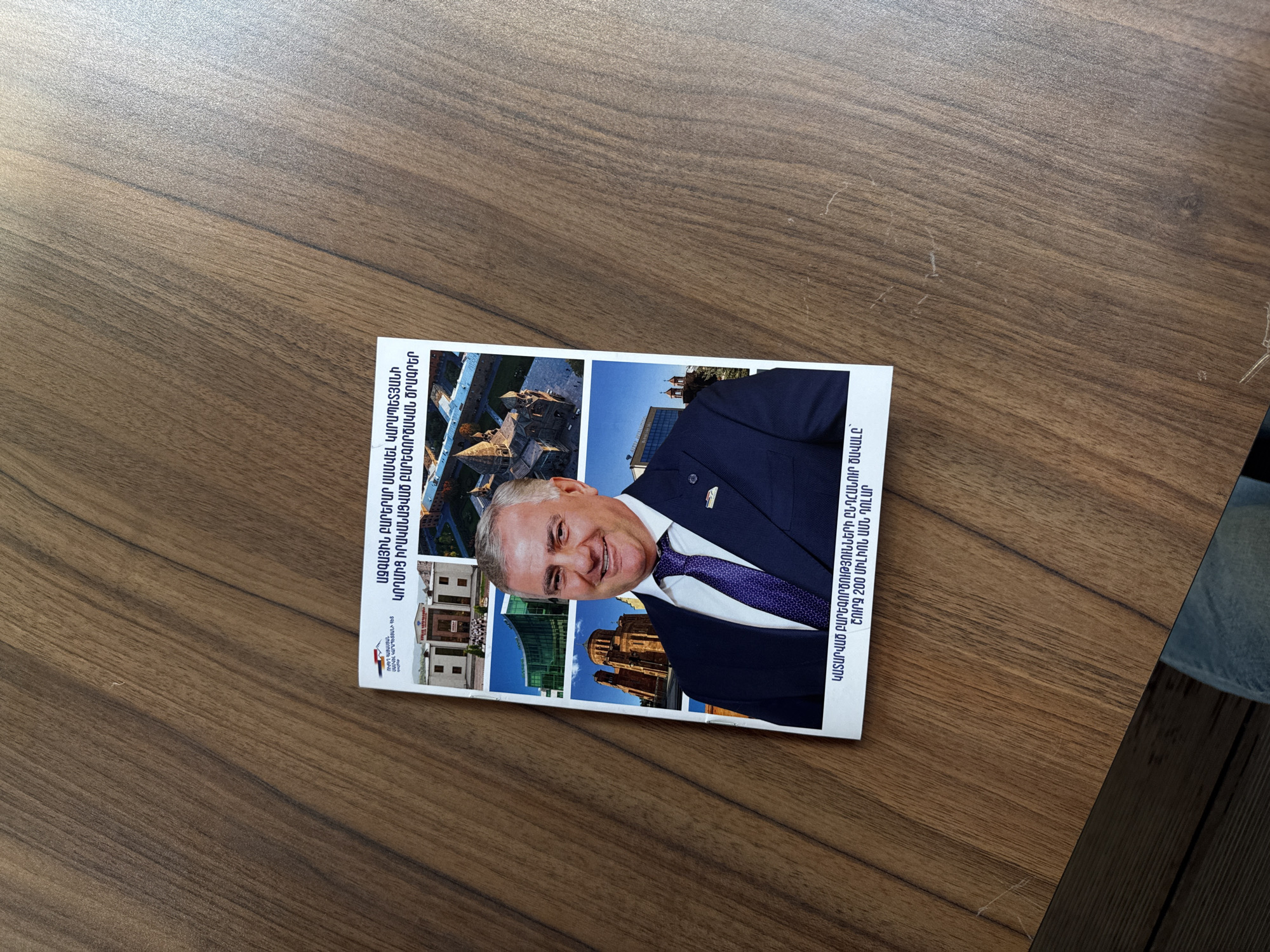 The image shows a booklet or pamphlet lying on a wooden surface, possibly a table. The booklet has Armenian text on it, and it features a portrait of a man in a suit and tie, along with images of buildings. The text mentions "National Benefactor Samvel Karapetyan" and "charitable programs." The text also mentions the total volume of charitable works is about 200 million US dollars. The location of the buildings is unclear from the image itself, though the text suggests they may be in Yerevan, Armenia, given the Armenian language and benefactor mentioned.