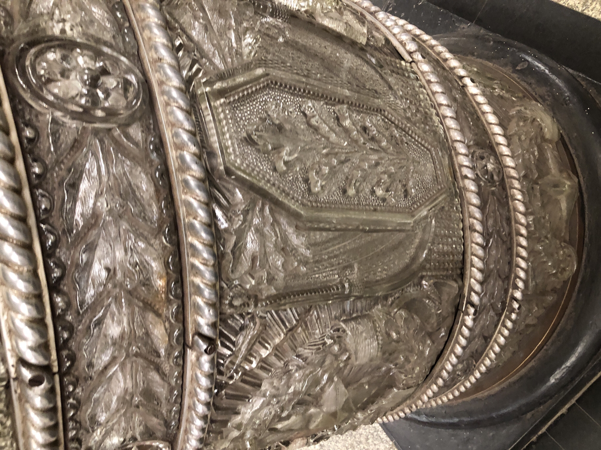 The image shows a close-up view of an ornate, possibly antique, decorative object. The object appears to be made of glass and metal, with intricate carvings and metallic trim that resembles rope. The glass sections feature detailed relief work, including what looks like floral patterns and possibly figural scenes. The metallic bands are twisted and textured, adding to the object's elaborate design. The object is situated on a dark, possibly stone or metal, base. The background is out of focus but appears to be a textured surface, possibly carpeting or a rough stone. There are no people or animals visible in the image. The lighting highlights the reflective surfaces of the object. The location in Saint Petersburg, Russia, is not discernible from the visual information provided in the image.