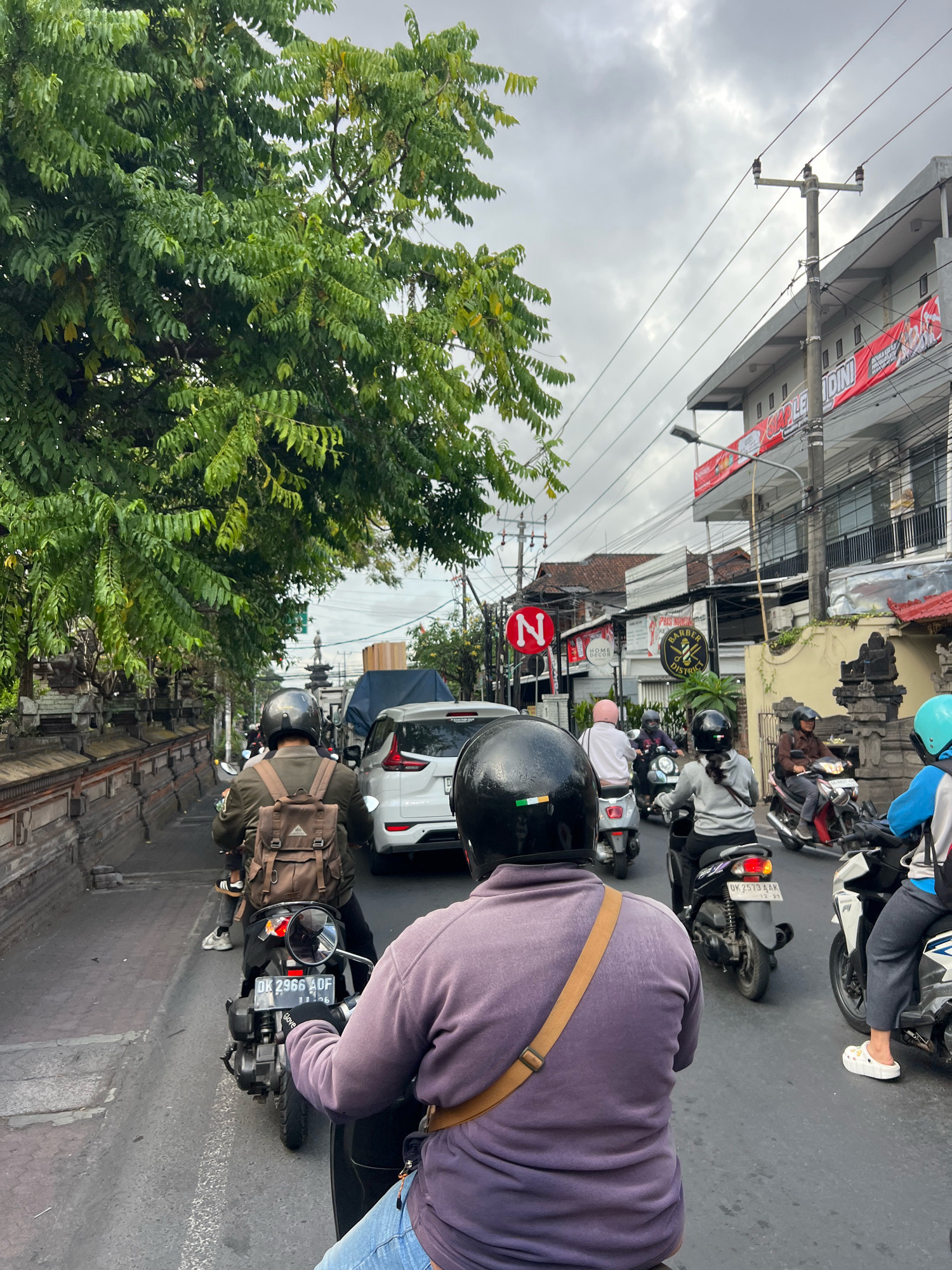 The image depicts a street scene in Denpasar, Indonesia, likely during a time of moderate traffic. A variety of motorcycles and a white car are visible on a paved road, some moving, others seemingly stopped. Several individuals are riding motorcycles, most wearing helmets. One individual in the foreground, viewed from behind, is wearing a purple long-sleeved shirt and blue jeans, with a brown backpack on their shoulders. Another person in the middle ground is also on a motorcycle, wearing a grey top and black pants, with their hair in a ponytail. The street is lined with buildings, some featuring banners with Indonesian text. Lush green trees overhang the street from the left side, and a stone wall with intricate carvings runs along the sidewalk. Power lines and utility poles are present overhead. The sky is overcast.