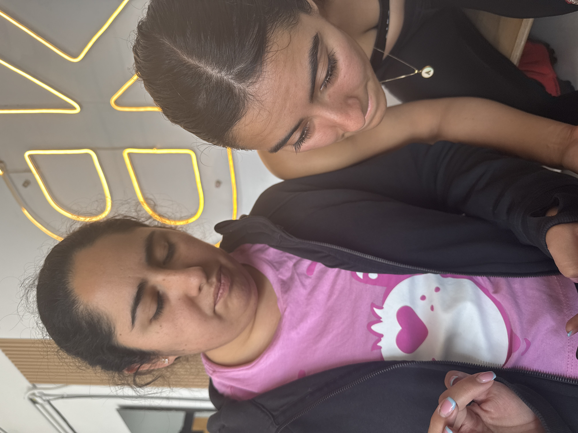 Two women are positioned close to one another. One woman is wearing a black top and a gold necklace with a white and gold pendant. The other woman is wearing a pink shirt with a white graphic featuring a pink heart. She also has a black zippered jacket over the pink shirt. The setting includes a bright yellow light fixture in the background. The specific location in Puebla, Mexico, is not identifiable from the given image.