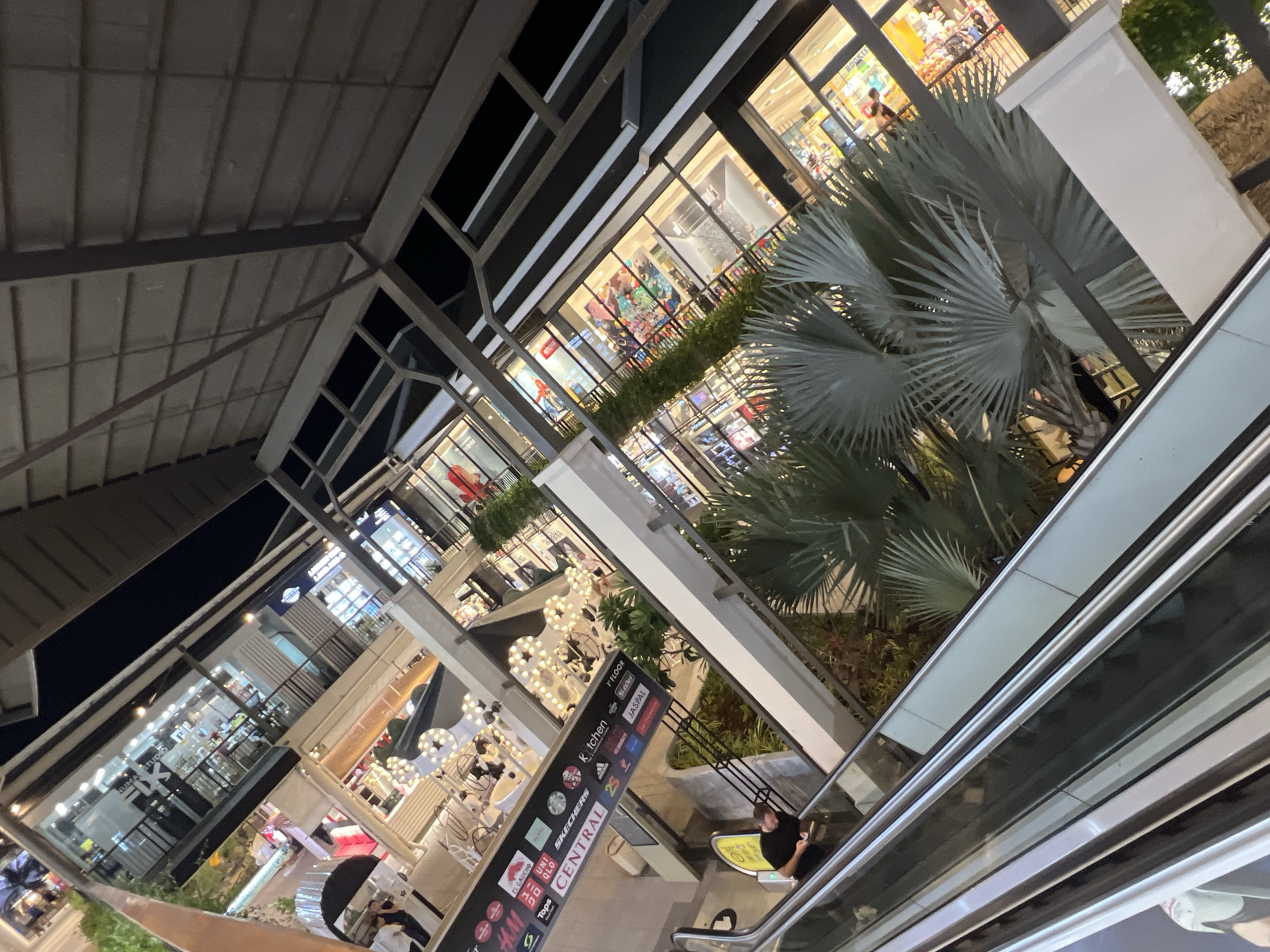 The image shows an outdoor shopping complex in the Chaweng area of Ko Samui District, Thailand. The perspective is from an escalator looking down and to the side. In the foreground, the handrail and moving steps of an escalator are visible. A person in a black shirt is standing on the escalator, looking at their phone. To the right of the escalator, a tall white pillar and a lush palm tree are present.
The middle ground of the image reveals the multi-level structure of the shopping complex. Shop fronts with large glass windows display merchandise and lighting. Various signage is visible, including a large sign that reads "CENTRAL" and lists brand names like "KITCHEN," "UNI QLO," "H&M," "SKECHERS," and "JASPAL." Decorative star-shaped lights are hung on some of the upper levels.
The background shows more of the shopping complex, with illuminated shop interiors and architecture that suggests a modern design. The sky is dark, indicating it is nighttime. The overall impression is a vibrant and active commercial area in Ko Samui.