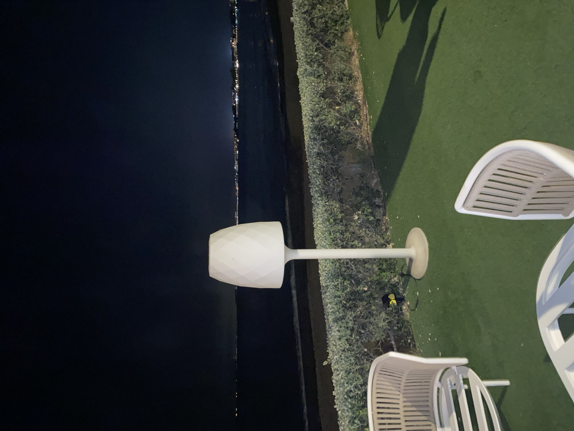 This is a nighttime, eye-level, full shot taken outdoors at a beachfront location in Estepona, Spain. The foreground features a patch of artificial green turf with shadows cast by unseen objects. Two light-colored, modern outdoor chairs with ribbed backs are partially visible on the turf. A white floor lamp with a diamond-patterned shade stands in the midground, casting a shadow on the turf. Behind the lamp is a low, manicured green hedge. Beyond the hedge is a dark beach area bordering a calm body of water, likely the Mediterranean Sea. In the distance, across the water, a line of lights indicates a coastal town or city skyline. The sky above is dark and clear, with a few faint lights visible in the upper left quadrant, possibly stars or distant aircraft. A small, yellow solar-powered garden light is visible near the base of the floor lamp.