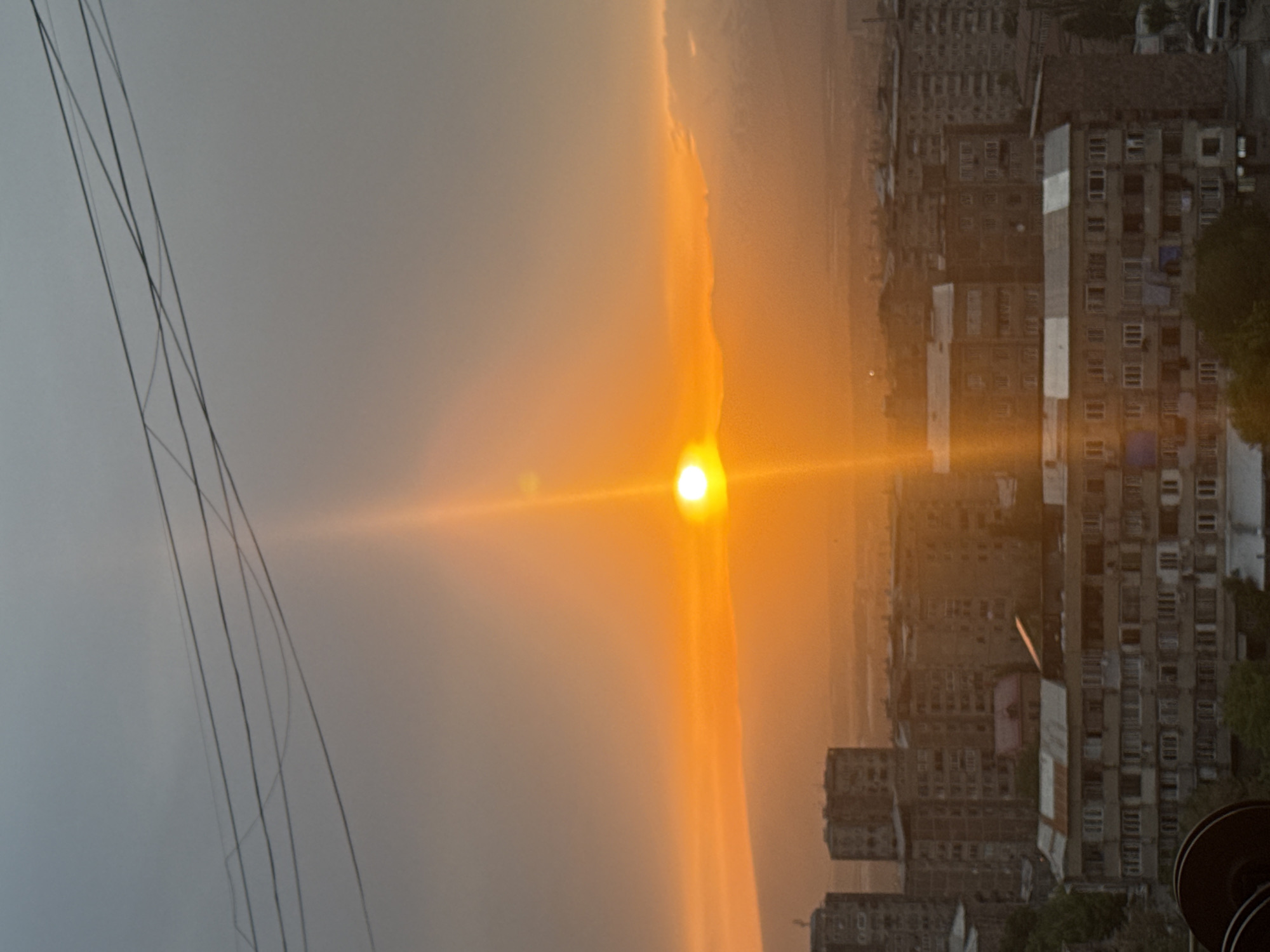 The image depicts a sunset in an urban environment, likely the city of Abovyan, Armenia. The sun is positioned prominently in the center, emitting a bright orange light that illuminates the surrounding sky. A layer of haze or smog diffuses the light. To the right, there is a view of densely packed apartment buildings, typical of urban residential areas. Power lines are visible on the left. The general ambiance is warm due to the colors of the sunset.