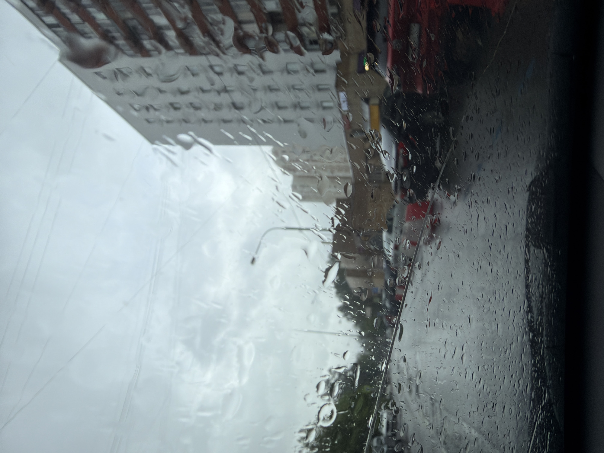 The image appears to be taken from inside a vehicle on a rainy day in an urban environment. Raindrops are heavily present on the glass, obscuring some of the view. In the background, one can discern buildings, including a tall building with multiple floors and a structured facade. There are hints of traffic and street infrastructure, such as a streetlight, though the details are blurred due to the rain on the window. The sky is overcast, contributing to the grey, muted tones of the scene. The vehicle's interior is suggested by the darker area on the right, indicating the edge of the window or dashboard.