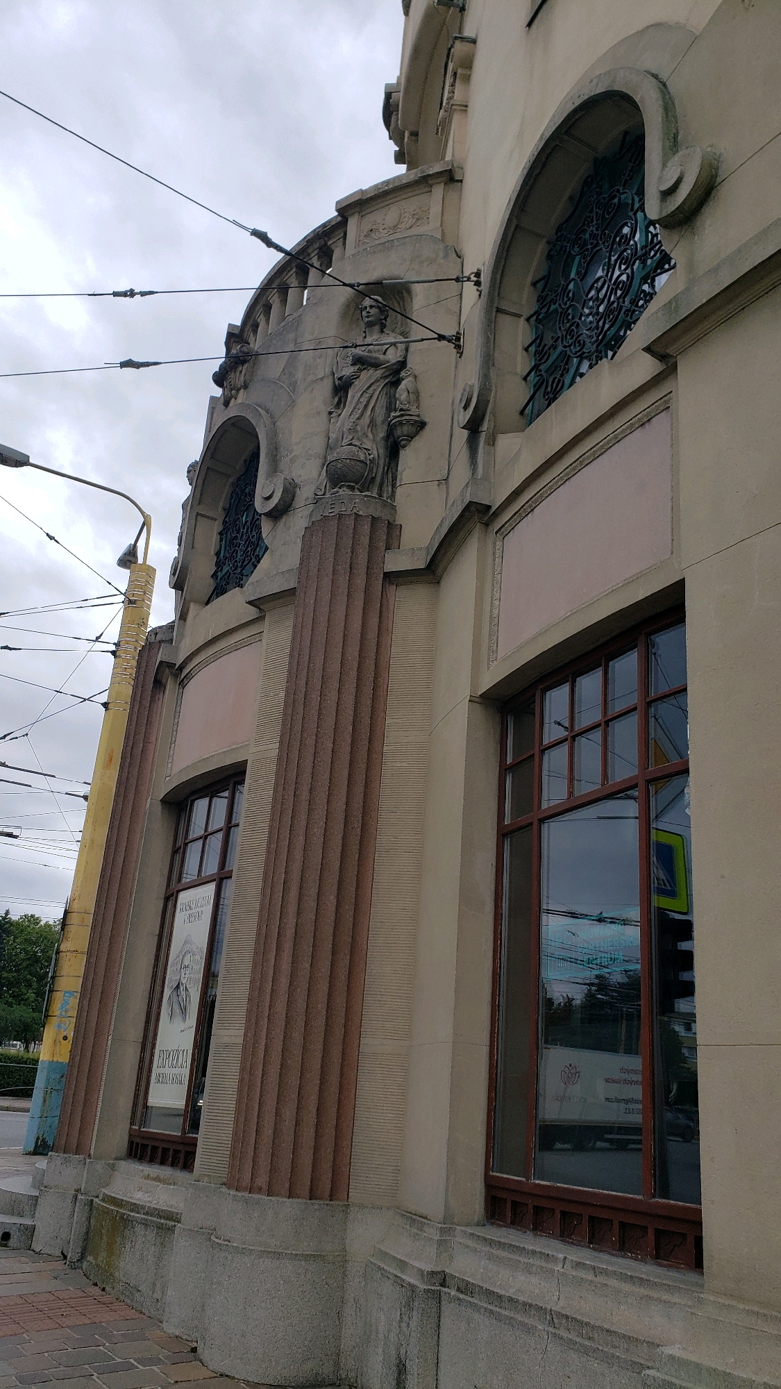 This is an exterior, low-angle shot of a building facade in Prešov, Slovakia, likely taken on an overcast day. The architecture appears to be from the early 20th century, with Art Nouveau or Art Deco influences. The building is made of light-colored stone and features decorative elements such as fluted columns with reddish-brown accents, arched windows with ornate metal grilles, and a stone sculpture of a draped female figure holding a lamp and resting her hand on a globe.
One prominent window on the lower left shows a poster with a portrait of a man and text that appears to advertise an exhibition. Reflections in the large window on the right reveal overhead power lines for a tram, trees, and possibly a traffic sign. The ground in the foreground is paved with brick. The overall impression is of a historic urban setting, likely a public building such as a museum or a cultural institution.