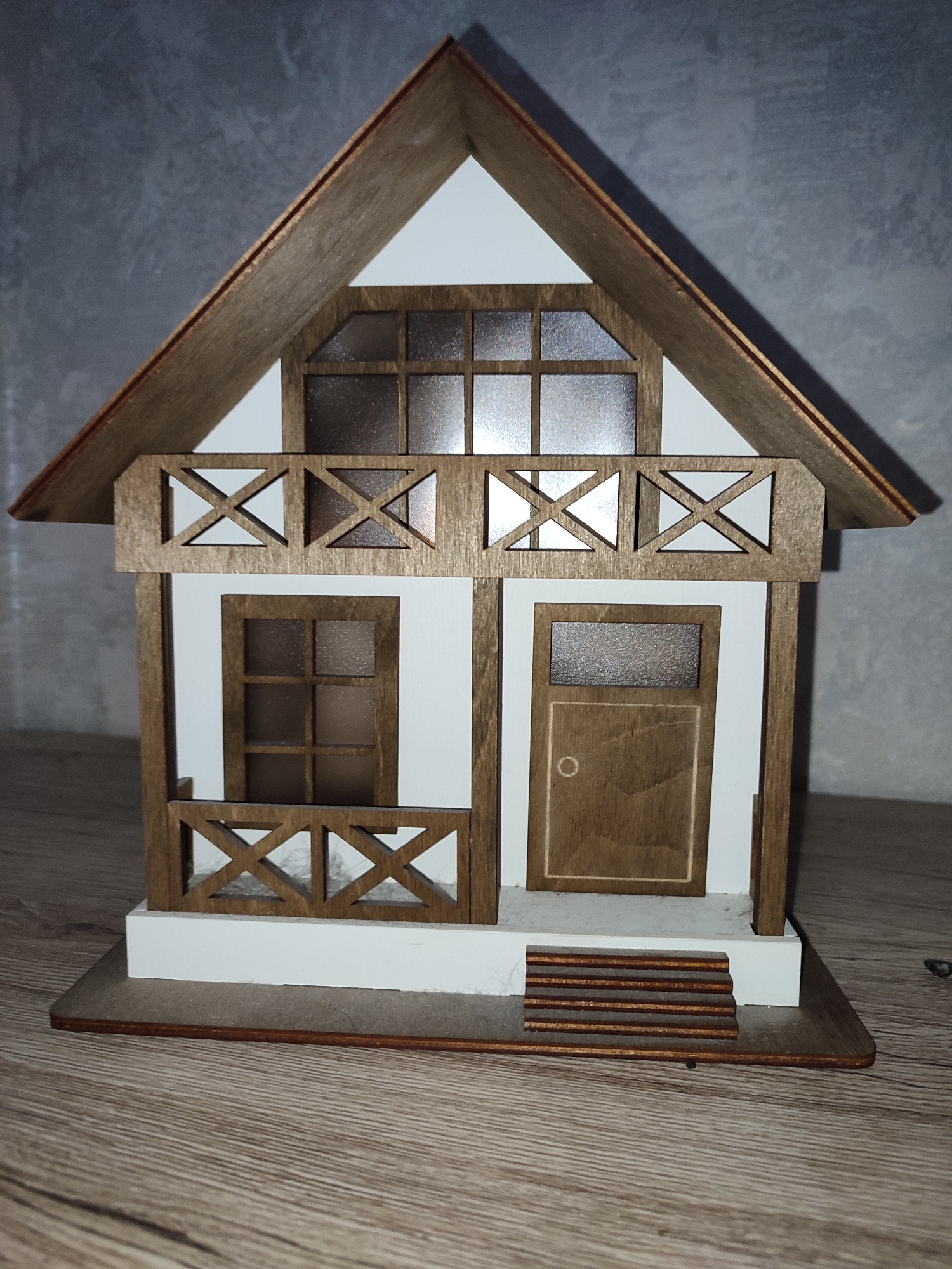 This is a close-up, eye-level, studio shot of a model house. The house is made of light-colored wood with white accents and features a pitched roof, a balcony with decorative wooden railings, a door with a round doorknob, and several windows. The windows have a frosted appearance and are divided into square panes by wooden mullions. A small set of wooden stairs leads up to the front door. The model house sits on a rectangular wooden base, which rests on a wooden surface. In the background, there is a gray, textured wall. The lighting appears to be artificial, and there are no people or animals present. The overall impression is of a quaint, rustic dwelling.