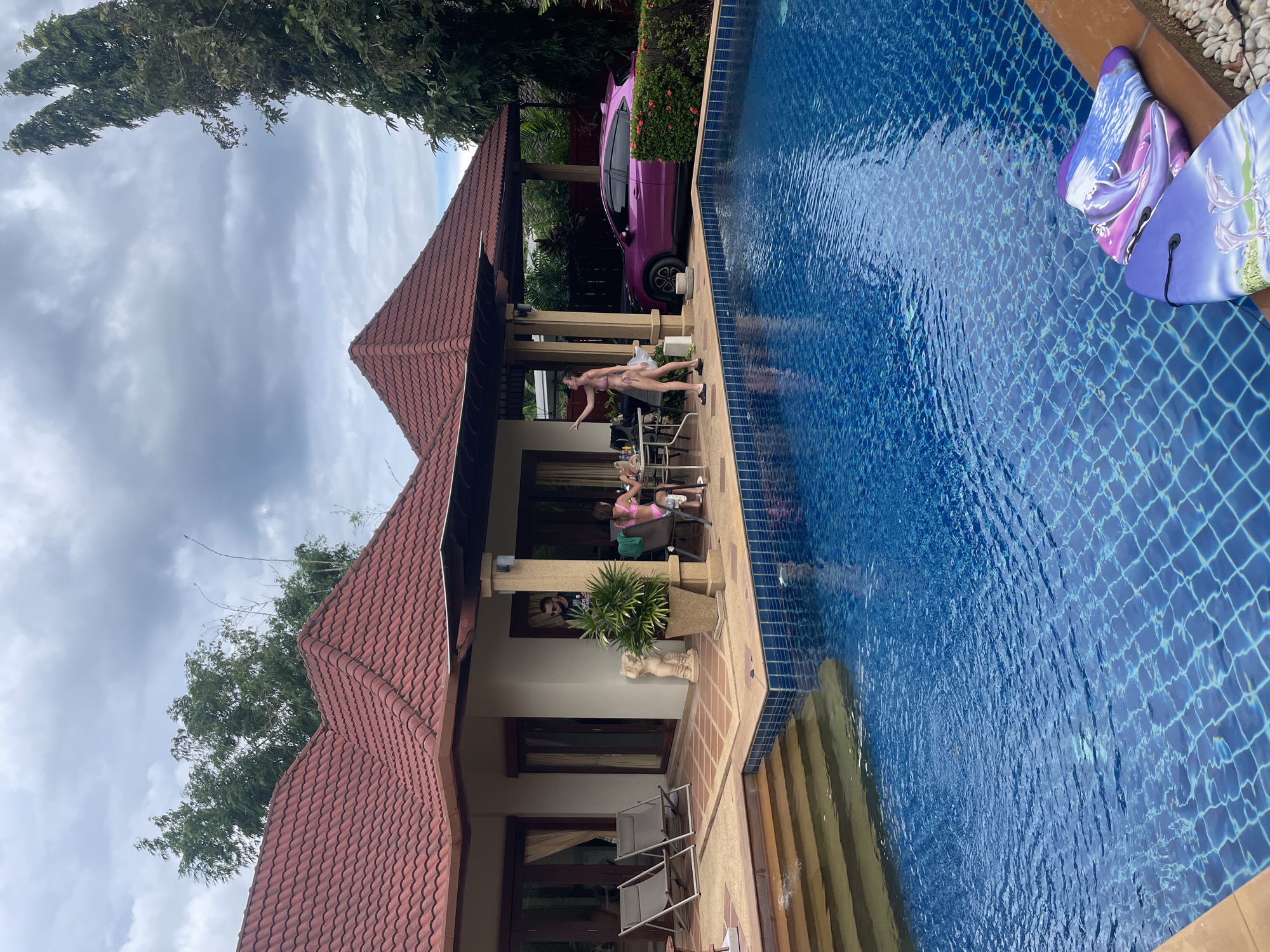 The image depicts a residential property with a swimming pool in Rawai, Thailand, under a cloudy sky. A house with a red tiled roof and an attached covered patio is visible on the left. Two lounge chairs are set up on the patio. Adjacent to the patio, a group of people are seated at a table, appearing to be dining or socializing. A woman in a pink bikini stands near the table, pointing towards the left. In the background, a bright pink car is parked under a separate covered area, next to a hedge with red flowers. The foreground features a large, blue tiled swimming pool with steps leading into it. Two boogie boards with dolphin and wave designs are resting on the edge of the pool. The overall scene suggests a relaxed, tropical vacation setting.
