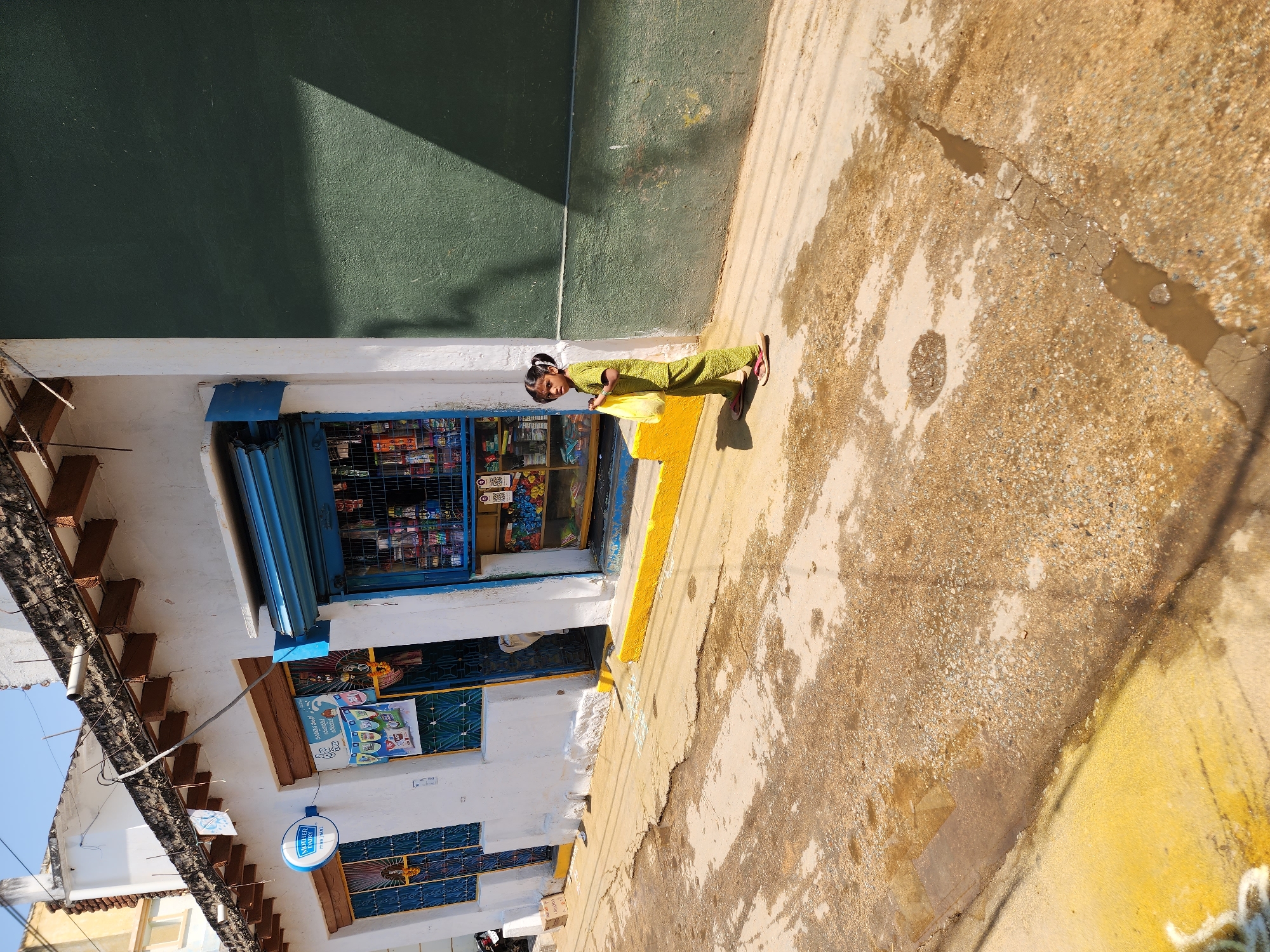 The image depicts a street scene in Pedda Kammavaripalli, India, under clear daylight with strong shadows. A young girl, approximately 5-7 years old, stands slightly right of center on an uneven, gravelly street surface. She wears a patterned light green two-piece outfit and light-colored flip-flops, with her dark hair in two pigtails. In her left hand, she holds a translucent yellow plastic bag. She is looking directly towards the viewer.
To her left, a white-painted building with a flat roof and exposed dark wooden ceiling joists features a blue "Mother Dairy" sign above a window with a blue grate. Adjacent to this, a shop entrance is partially open, revealing a blue rolling shutter above shelves stocked with various packaged goods. A yellow painted curb runs along the base of this building. The ground in front of the shop shows white chalk-like markings. Further left, another blue-grated window and a small section of a motorcycle are visible.
To the girl's right, a plain, dark green textured wall stands, with a distinct triangular shadow cast across its upper portion. The ground beneath the girl and extending into the foreground is cracked and features several darker, wet patches, including a narrow puddle in a crack extending towards the upper right. The sky overhead is clear blue.