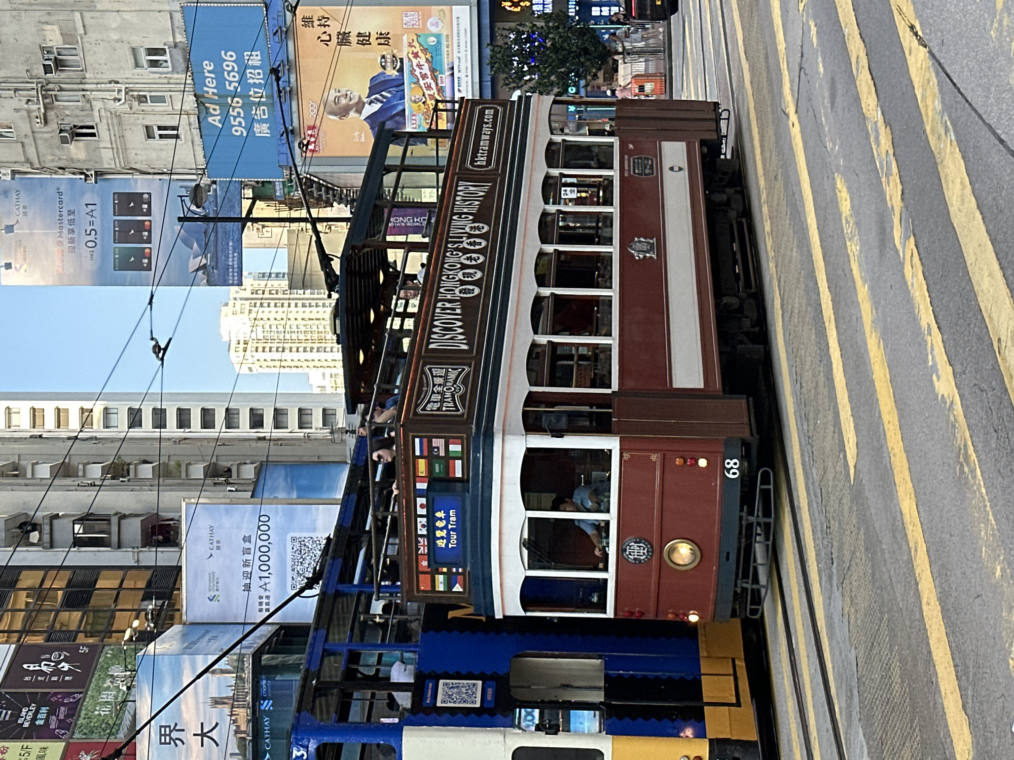 In this medium-shot, street-level photograph, a vintage Hong Kong tram, painted maroon and white with a brown roof, travels on a city street. The tram is numbered 68 and displays flags of various countries along its upper deck, along with the text "Tour Tram" in Chinese and English. Another tram, predominantly blue and yellow, is visible to the left. The street is marked with yellow pedestrian crossing lines. In the background, tall buildings with multiple windows and advertising billboards are present, including one for Cathay Pacific and Standard Chartered. The sky is clear and blue. A few individuals are visible on the upper deck of the maroon tram, appearing to be tourists. The overall scene captures a typical urban environment in Hong Kong.