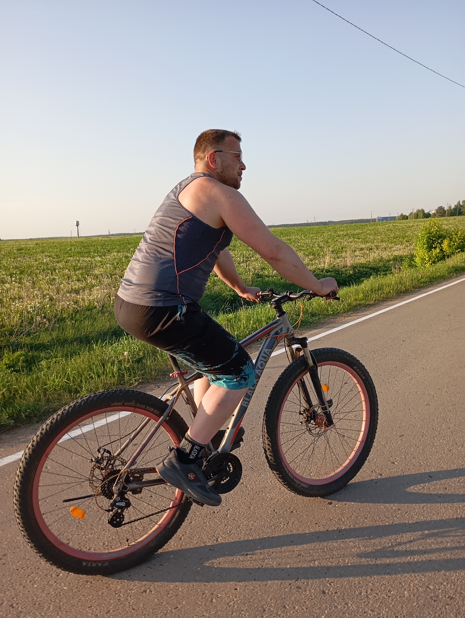 A person is riding a bicycle on a paved road. The person is wearing a gray tank top with black accents, black shorts, and gray athletic shoes with black socks. The bicycle is a mountain bike with pink rims. The background consists of a green field on one side of the road and a road on the other side. The location is Unknown, Unknown.