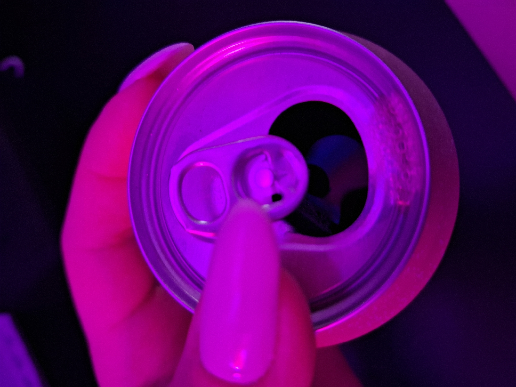 A close-up, overhead image depicts a person's hand holding an open metallic beverage can. The index finger, featuring a glossy light pink or purple oval-shaped nail, is positioned adjacent to the can's circular opening. The can's pull tab is depressed inward, partially obscuring the dark interior where subtle liquid residue and small bubbles are visible along the inner rim. The entire scene is bathed in intense magenta and purple lighting, creating a high-contrast environment against a dark, out-of-focus background. The specific geographical location, including the city of Zemun Urban Municipality, Serbia, is not discernible from the visual content.