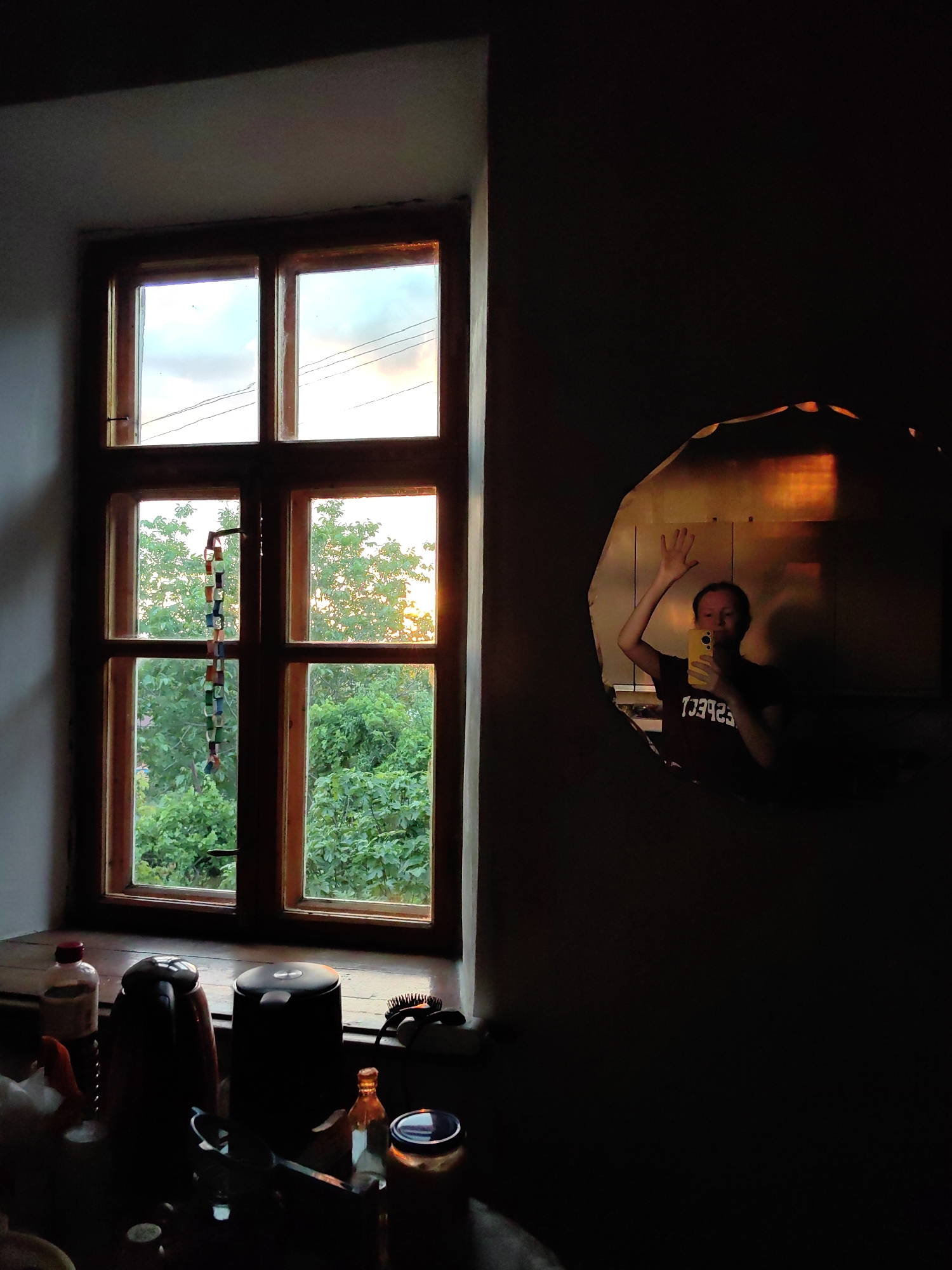 The image depicts an indoor scene with a wooden-framed window on the left, revealing a lush green outdoor environment with trees and a cloudy sky. A string of colorful beads hangs from the window. On the windowsill, several objects are visible, including bottles, kettles, and a hairbrush. To the right of the window, a person is reflected in an irregularly shaped mirror, holding a yellow smartphone and waving. The mirror is positioned on a dark surface, and the reflection shows the person wearing a dark t-shirt with text that appears to read "ESPECT" in reverse. The overall lighting suggests it is either early morning or late afternoon, with warm light filtering through the window and reflecting in the mirror. The setting is likely a room within a dwelling in Зуйское сельское поселение, Ukraine.