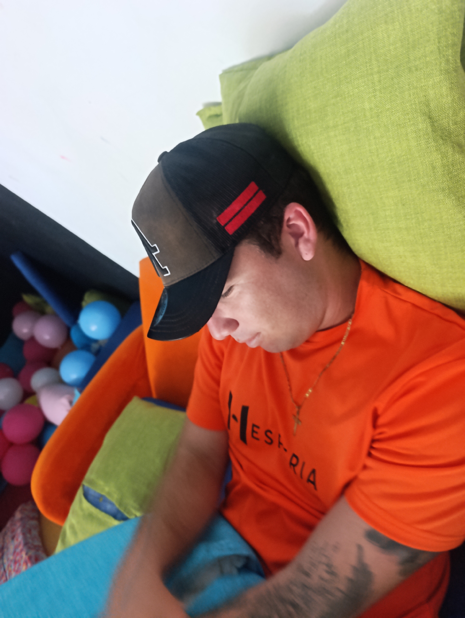 A man is resting in a colorful environment. He wears a black and brown baseball cap, an orange t-shirt with lettering, and a gold necklace with a cross pendant. His arm has a tattoo. He leans against a lime green cushion. An orange chair with a green cushion is visible, along with a ball pit with colorful balls. The location is in the city of Unknown, Unknown.