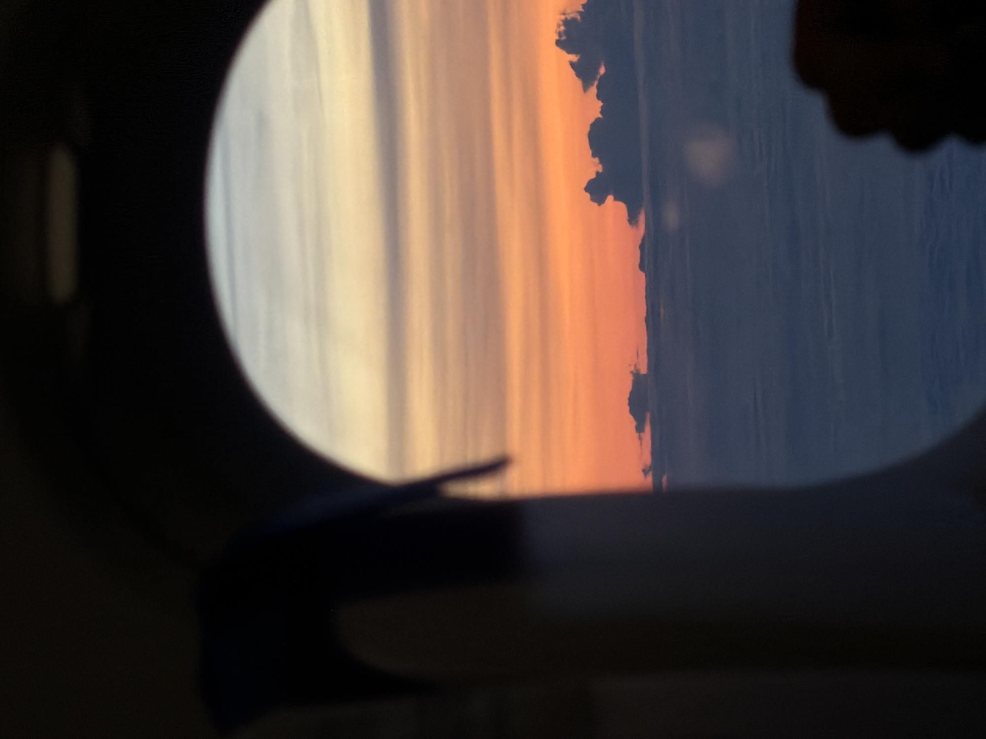 A view from an airplane window shows a vibrant sunset over a cloud-covered landscape. The sky displays a gradient of colors, transitioning from pale yellow and white at the top to warm oranges and pinks closer to the horizon. Dark, wispy clouds are scattered above a thicker layer of clouds that stretches across the lower half of the frame. The bottom right corner shows the silhouette of a hand in the foreground. The location is not specified to be in Nadergul, India.