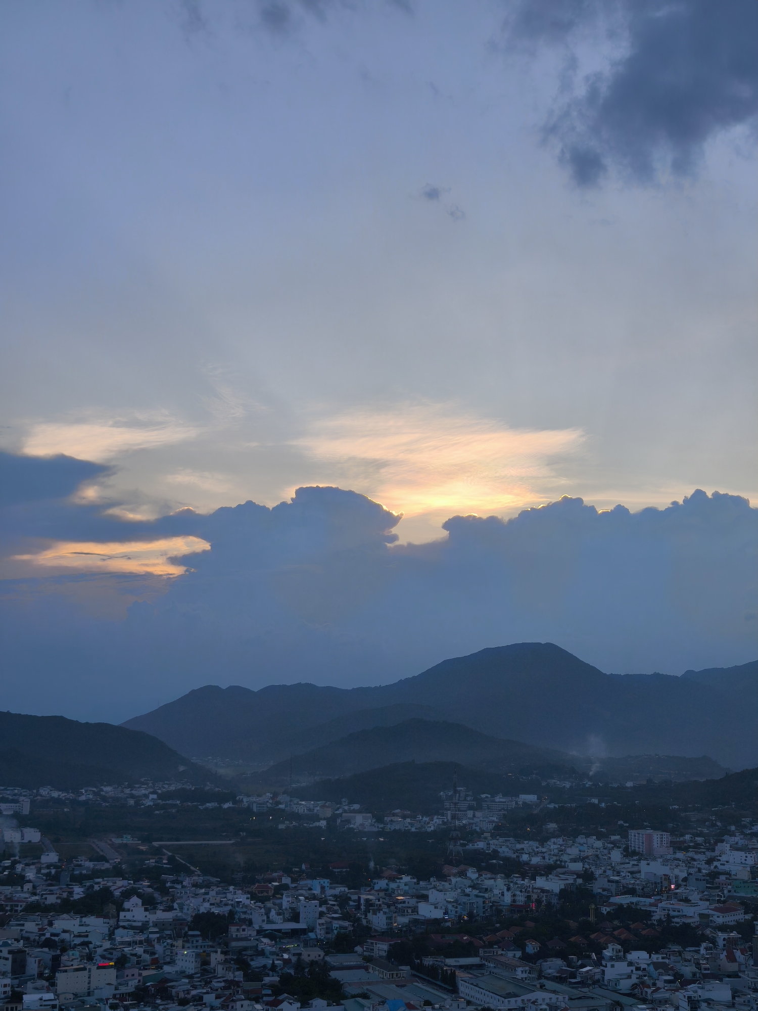 The image depicts an aerial view of a city at dusk, likely Bac Nha Trang Ward, Vietnam. Rolling hills and mountains form the backdrop, their slopes partially obscured by the fading light and atmospheric haze. Nestled in the valleys and plains between the hills is a densely populated urban area. Numerous buildings, primarily residential with varying architectural styles and colors, cover the landscape. Some structures are taller and more modern, while others appear to be older, with tiled roofs. Streets and smaller roads weave through the city, punctuated by patches of greenery and trees. A faint glow emanates from some of the buildings, suggesting that lights are beginning to turn on as night approaches. The sky above is a blend of twilight hues, with dark, heavy clouds dominating the upper portion and lighter, warmer tones of orange and yellow appearing near the horizon, hinting at the lingering glow of the setting sun. The overall impression is one of a peaceful, developing urban landscape transitioning into evening.