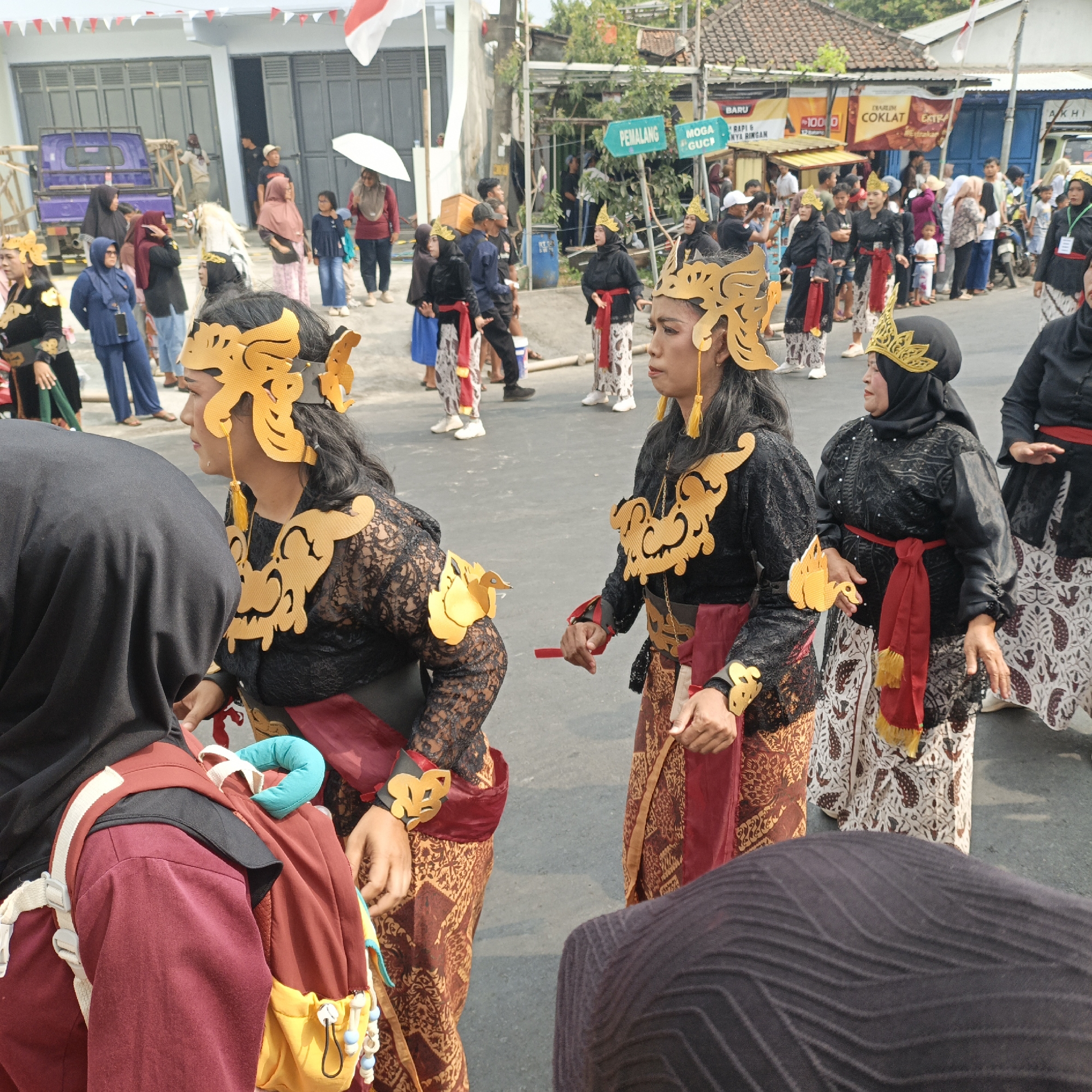 In Randudongkal, Indonesia, on a paved road lined with buildings, a group of people are participating in a parade or cultural event. The foreground features several individuals dressed in traditional Indonesian attire. One woman in the center wears a black lace top with gold decorative elements on her shoulders and chest, a red sash tied around her waist, and a patterned batik skirt. She has a gold headdress on her head. To her left, another woman, also in similar traditional clothing, faces away from the camera. In the background, other participants in traditional dress are visible, along with spectators and some vehicles. The environment suggests a daytime outdoor event with a festive atmosphere.