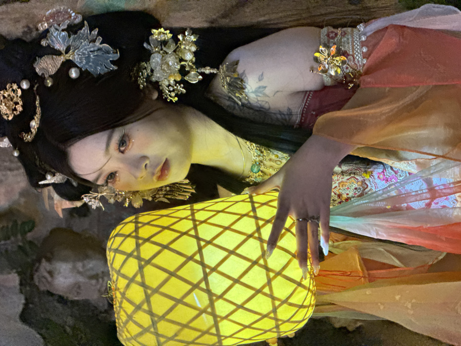 This image captures a woman in traditional Chinese-inspired attire, dramatically lit by a glowing lantern. She is positioned with her head turned, looking towards the viewer with a serene or contemplative expression. Her dark hair is elaborately styled with numerous ornate golden, silver, and pearl hairpins, featuring floral and leaf motifs. Her makeup includes subtle glitter on her eyelids, enhancing a delicate aesthetic.
The woman wears a richly embroidered garment with patterns in red, pink, and green, adorned with a golden arm cuff and a necklace featuring a prominent green gemstone. Flowing sheer fabrics in shades of orange, pink, and light blue are draped around her, adding to the ethereal quality of the scene. A dark tattoo is visible on her left arm, peeking from beneath her sleeve. Her hand, adorned with a ring and long, light-colored nails, rests gently on a large, cylindrical yellow lantern. The lantern, featuring a distinct cross-hatch pattern, is the primary light source, casting a warm, bright glow on her face, hand, and parts of her clothing.
The background is dimly lit and features dark, rough textures, possibly rocks or foliage, suggesting an outdoor or garden setting at night. The overall atmosphere is mystical and elegant, indicative of a staged photoshoot with a strong cultural or historical theme in Yuzhong District, China. No text is visible in the image.