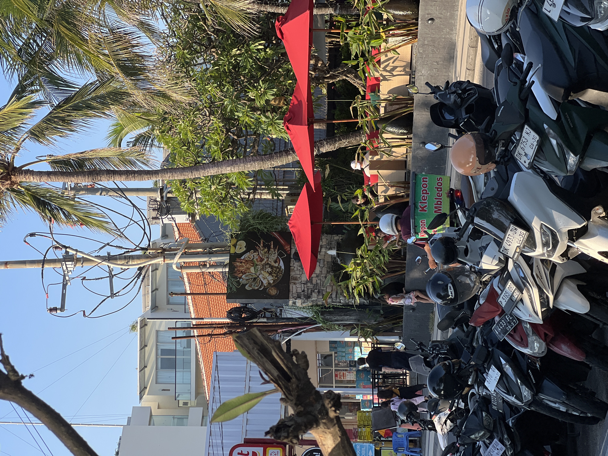 The image captures a street scene in Kuta, Indonesia, during daylight. A row of parked motorcycles, mostly scooters, occupies the foreground. Several of these scooters are adorned with helmets. In the mid-ground, a restaurant or cafe area is visible, featuring red umbrellas providing shade over outdoor seating. Patrons are seated at tables, some of whom appear to be interacting or dining. Lush green foliage and palm trees are abundant, creating a tropical ambiance. A large sign depicting seafood is displayed on a stone-clad wall behind the seating area. In the background, modern buildings and utility poles with numerous wires are discernible against a clear blue sky. The overall impression is that of a bustling street scene in a popular tourist area.