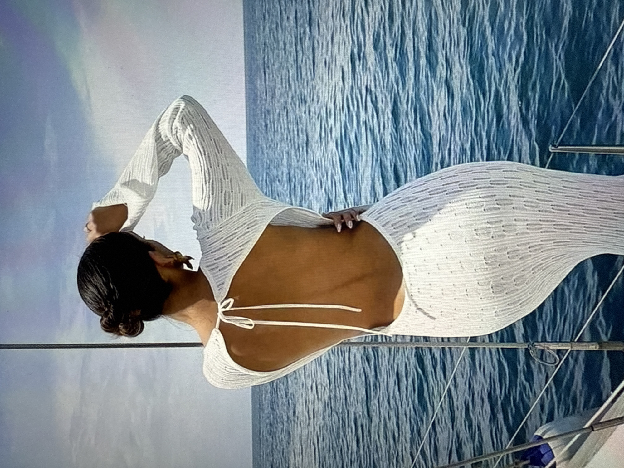 A woman with dark hair pulled into a bun, wearing a long-sleeved white dress with a low back and a cutout, stands on the deck of a boat. She is facing away from the camera, looking out at the blue ocean. Her right hand is raised to her head, and her left hand is resting on her hip. The dress appears to be made of a textured knit fabric. The boat is partially visible in the foreground. The sky is a pale blue with some clouds. The image does not provide any specific details about the location being in Găvănoasa, Moldova.