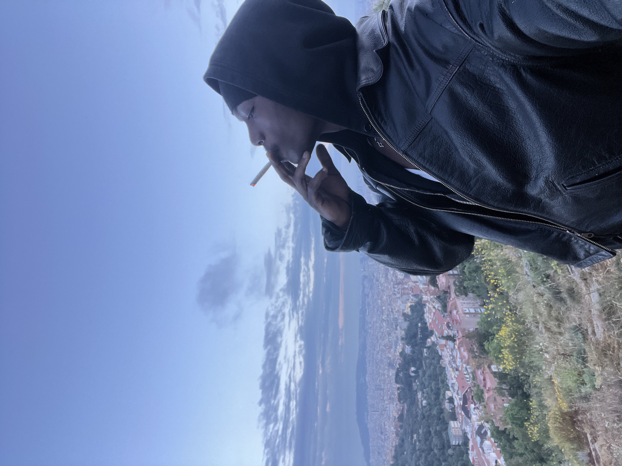 A man in a black leather jacket and hooded top stands facing left while smoking a cigarette. He is on an elevated area overlooking a sprawling cityscape and a body of water. The city is Barcelona, Spain. The photo is taken from a low angle, emphasizing the height of the man's vantage point. The sky is visible in the background.