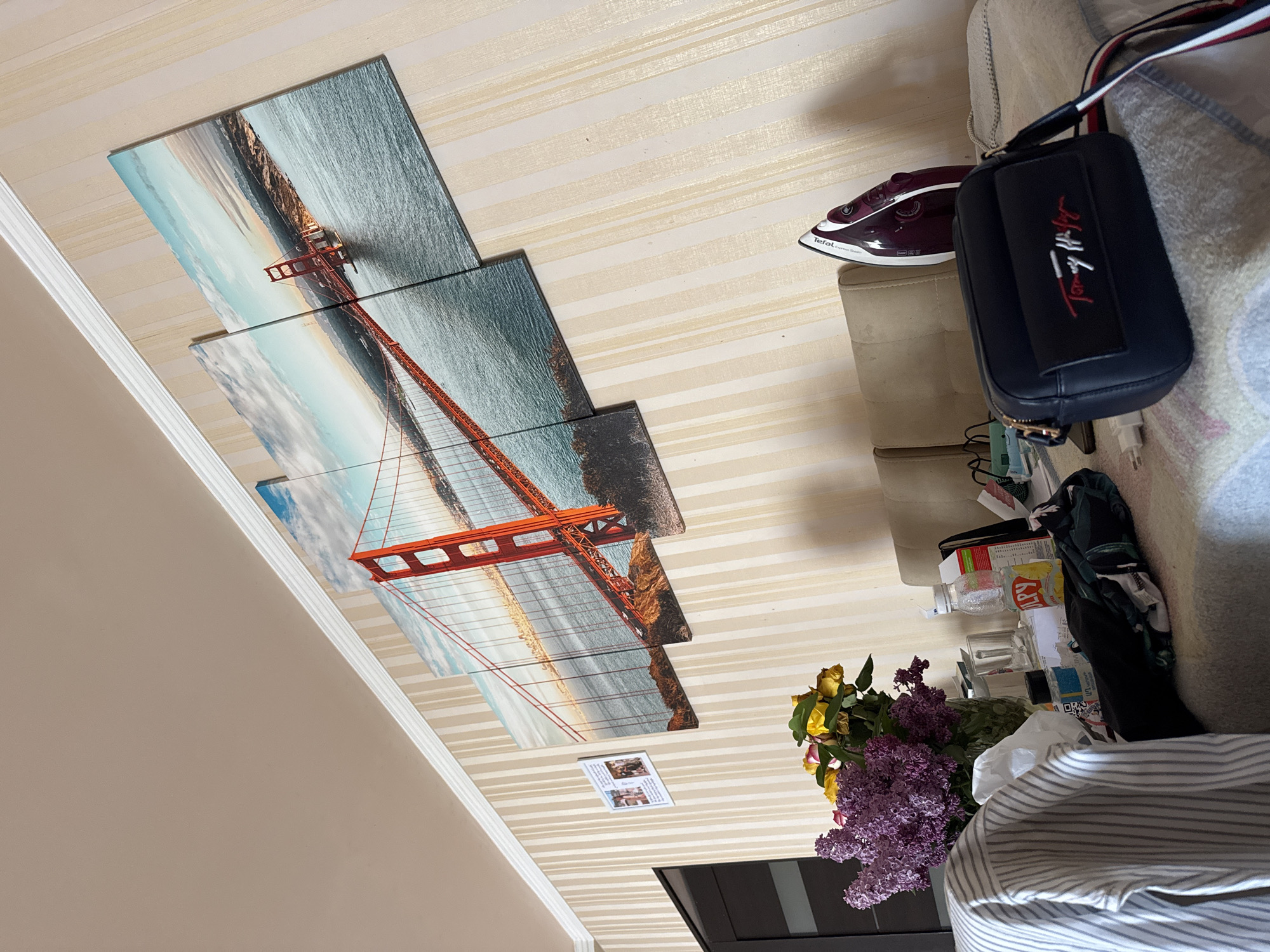The image, rotated 90 degrees clockwise, captures an indoor setting, likely a room in a home in Yerevan, Armenia.
The dominant feature is a wall adorned with striped wallpaper, featuring alternating bands of light beige and a slightly darker, textured beige. On this wall, a large, multi-panel artwork depicts the Golden Gate Bridge. The artwork is composed of at least four vertical segments, showcasing the iconic red structure against a backdrop of blue water and a sky with scattered clouds. A smaller, white rectangular card, displaying several miniature, blurred photographs and unreadable text, is also affixed to the wall below the bridge artwork.
To the right of the frame, a beige upholstered sofa or armchair is visible. A maroon-colored Tefal Express Steam iron rests on its arm or a nearby surface. Adjacent to this, a navy blue Tommy Hilfiger shoulder bag is prominently displayed. The bag features a distinct "Tommy Hilfiger" logo in red and white on a black patch, and its strap is decorated with red, white, and blue stripes.
Below these items, a cluttered surface holds a bouquet of flowers, including vibrant purple lilacs and several yellow roses, some of which appear slightly wilted, arranged in a clear vase. Other items on this surface include a plastic bottle with "LAY'S" visible on its label, various small clear bottles or containers, papers, and a white power adapter plugged into an outlet. A portion of a grey and white striped shirt is also visible in the foreground. No people are present in the image.