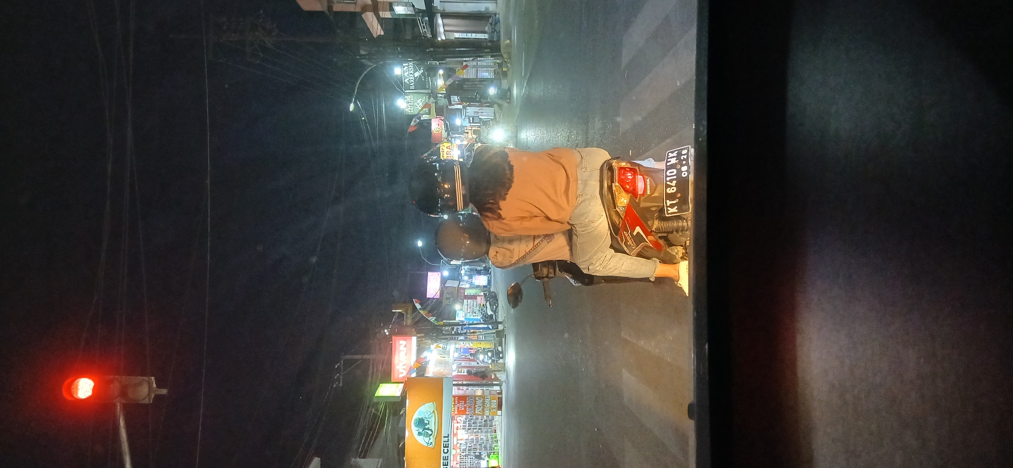 The image depicts a street scene at night in Samarinda, Indonesia, likely at an intersection with a traffic light. The traffic light is red, indicating that vehicles should stop. In the foreground, two people are riding on a motorcycle, facing away from the camera. They are wearing helmets, and the person in the back is wearing an orange sweatshirt and jeans. The motorcycle has a license plate that reads "KT 6410 WK".
The background shows a street lined with shops, some of which have illuminated signs. One sign on the left reads "BEE CELL". Streetlights and the lights from the shops illuminate the scene. There are power lines overhead. The road appears wet, suggesting it may have rained recently or the street is being cleaned. The overall impression is a typical urban night scene in Indonesia.