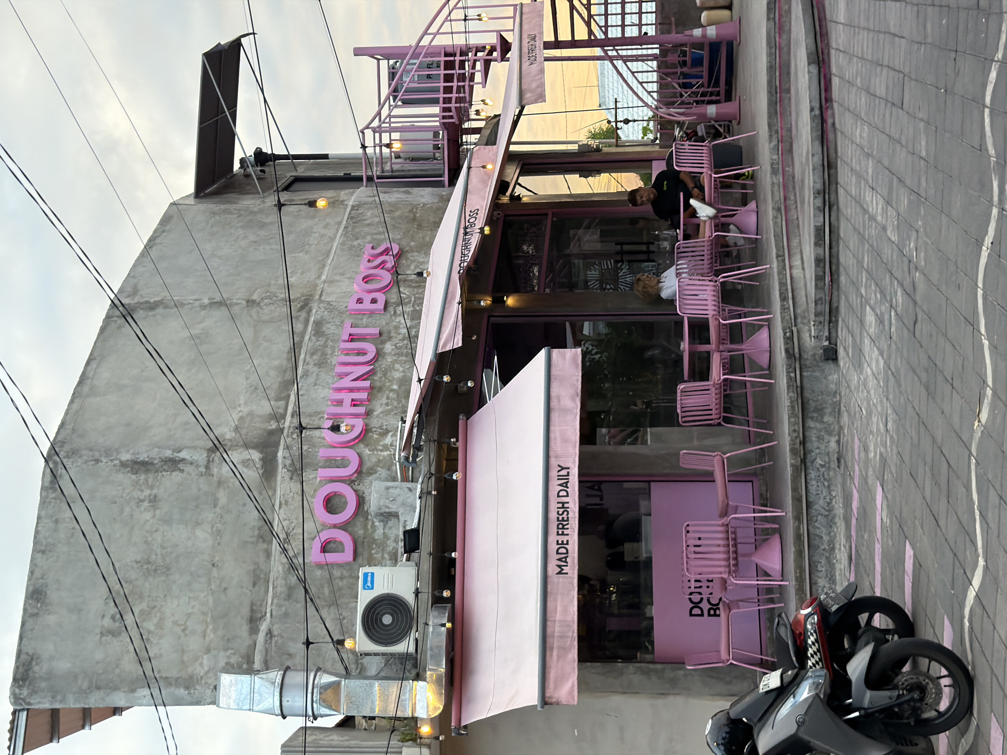 This image shows the exterior of a cafe called "DOUGHNUT BOSS" located in North Kuta, Indonesia. The cafe has a modern industrial aesthetic with a rough concrete facade and exposed metal piping. The cafe prominently features pink accents, including a retractable awning, outdoor seating, and a spiral staircase. Pink metal chairs and tables are arranged on the patio. Two people are seated at one of the tables, a woman with blonde hair wearing a white shirt and a man in a black t-shirt. In front of the cafe, two scooters are parked on the paved sidewalk. The sky is overcast, suggesting it is either early morning or late afternoon.